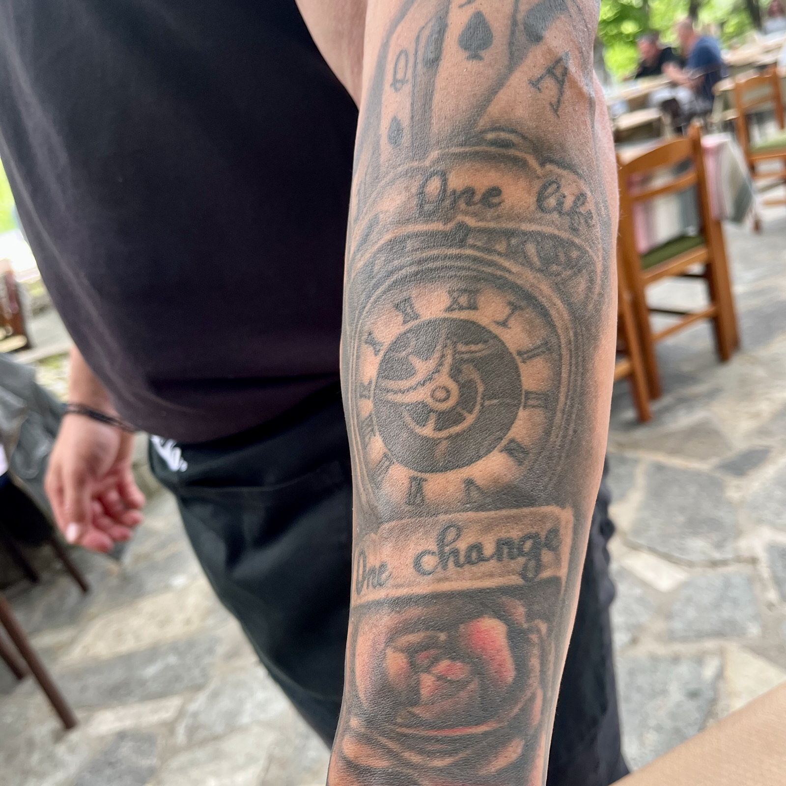 A large tattoo on someone’s underarm. It has playing cards, a rose, a clock, and the words One Life, One Change on it. The clock has no hands, so it doesn’t tell us what time it is.