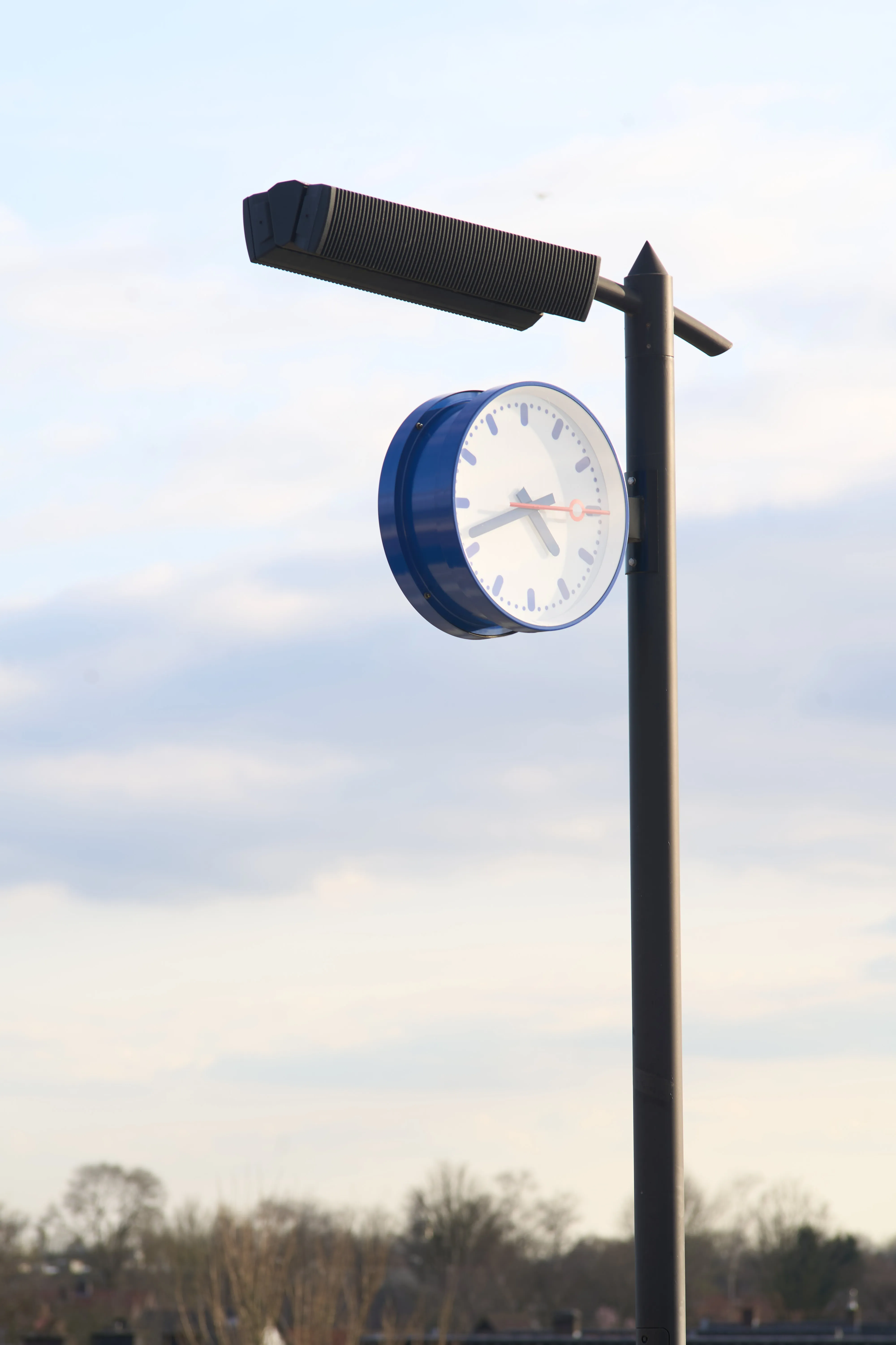 A station clock attached to a lamp post, somewhere in a rural part of the Netherlands. It points at 19 minutes to 5.