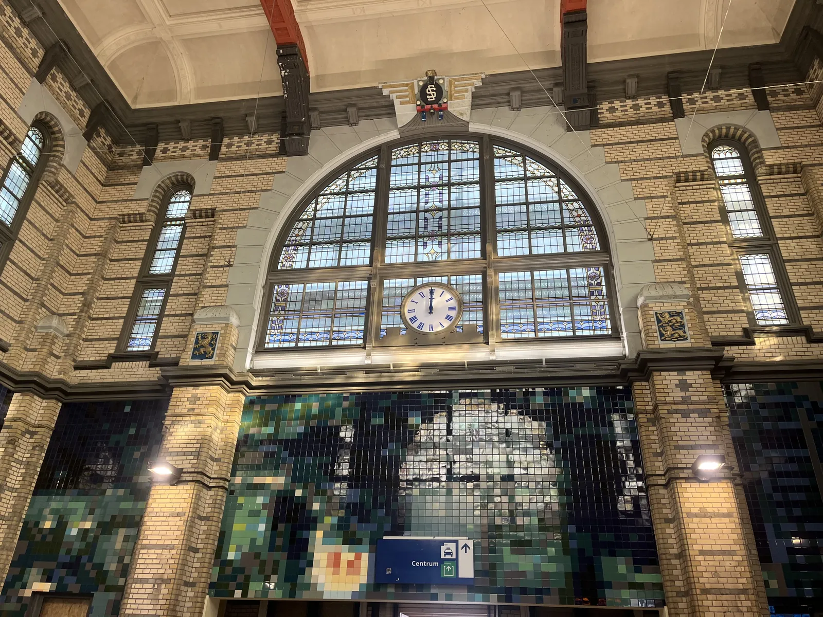 A clock built into the window of a Dutch train station. It’s an eclectic building. The windows, the roof, and many ornaments are old, while the large mosaic is clearly from a more recent date. Both hands of the clock point straight up, it is 12 o’clock.
