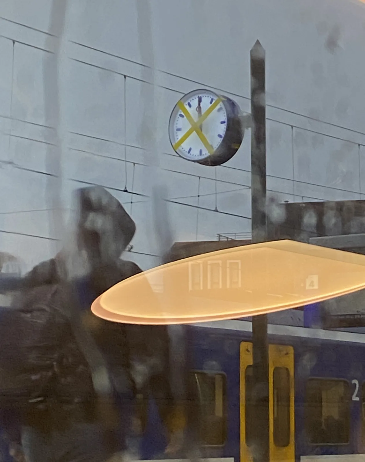 Looking out a window of a train. There’s some reflection. Outside it’s raining. There’s a clock on a pole with an X taped over it. Behind the X you can see that the clock points at exactly twelve o’clock.
