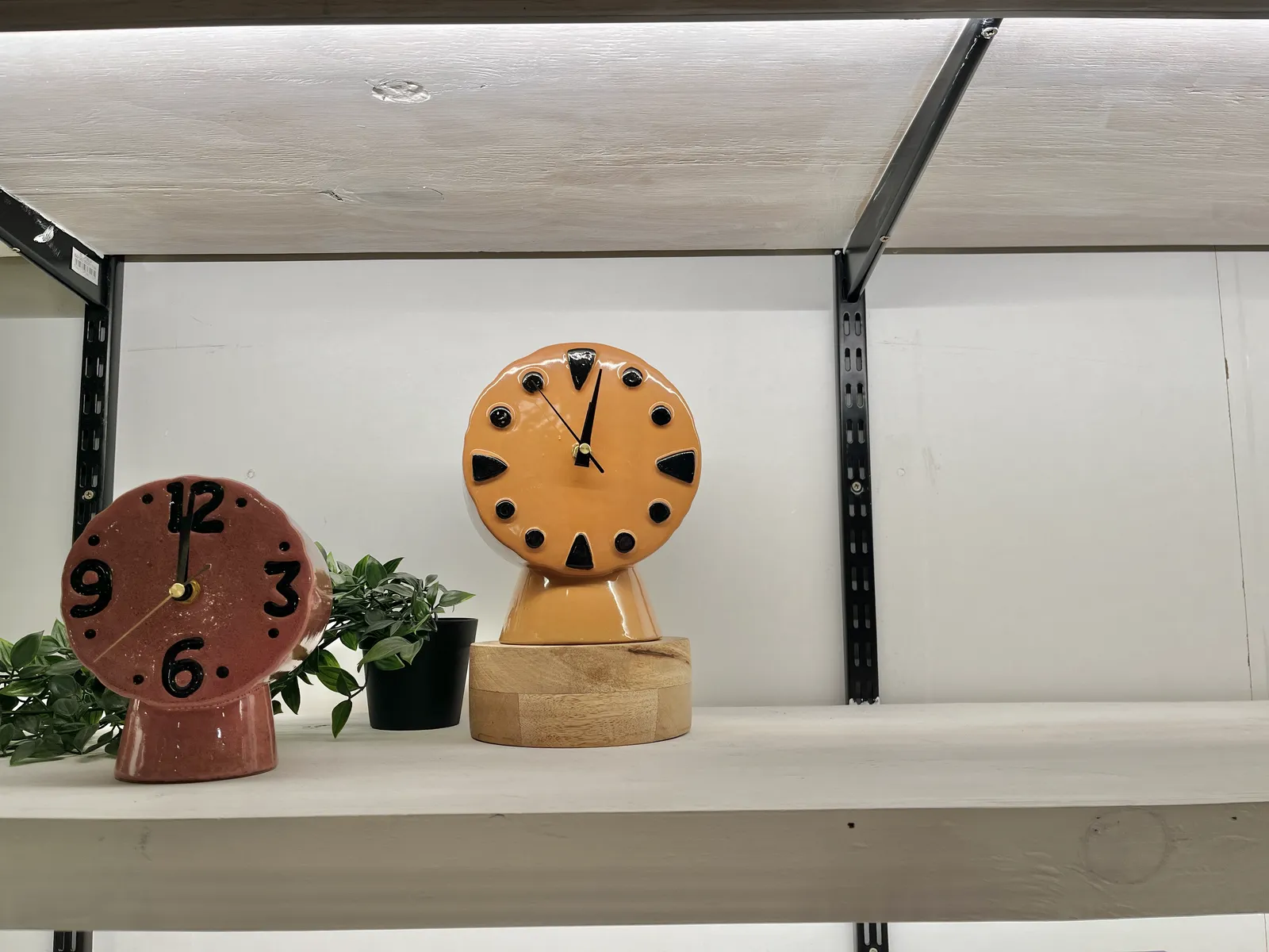 Two ceramic clocks on a shelf. The one on the left, the red one, points at twelve o’clock, the yellow one points at two minutes past twelve.