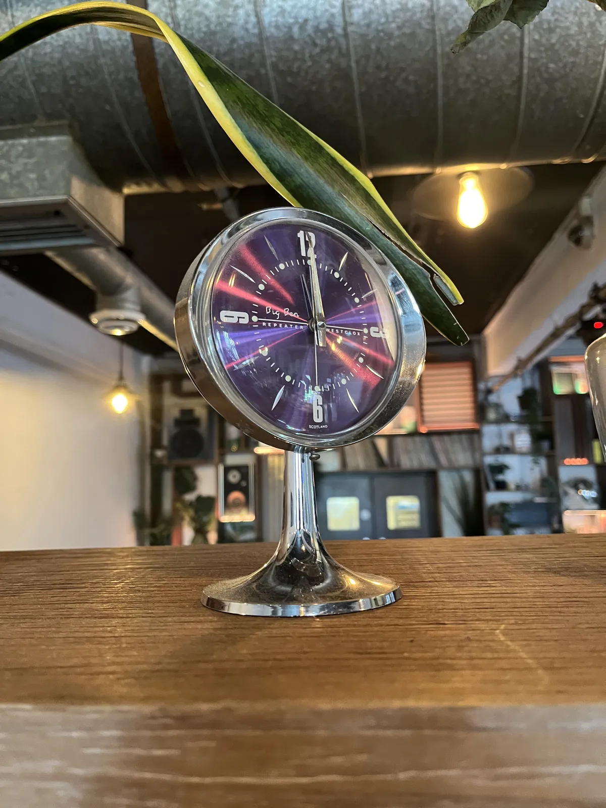 The background of this image is blurry, but a bookcase can be distinguished. In the foreground there’s a modern silver clock on a silver point with a dark blue, almost purple face. It points at twelve o’clock.