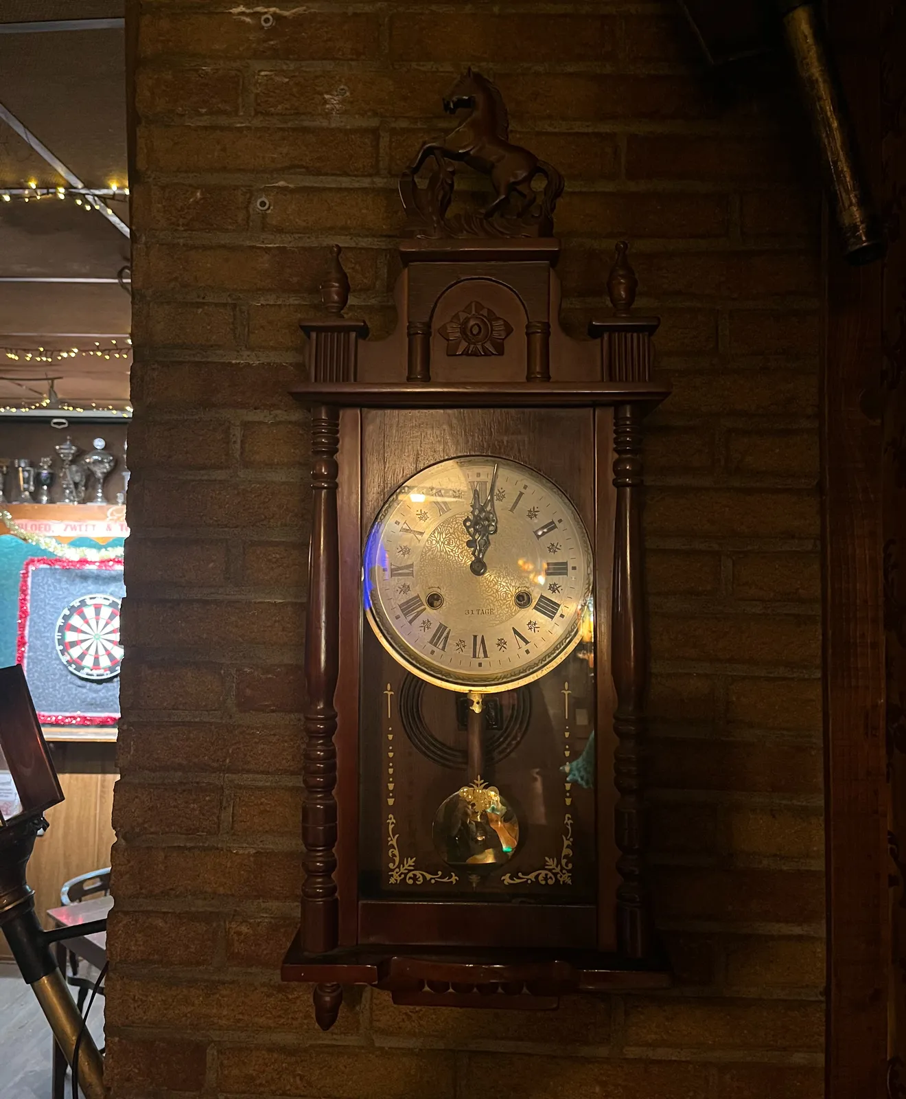 A wooden clock in a bar with a little horse on top. It points at one minute past 12.
