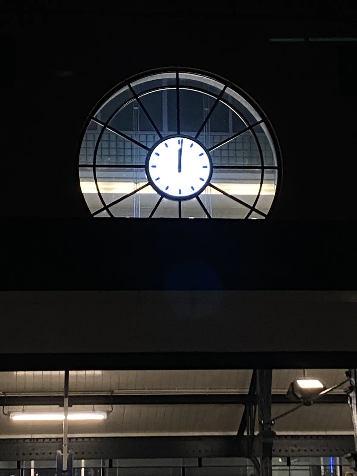 A modern clock, fitted in a round window at night. It points at one minute past twelve o’clock.