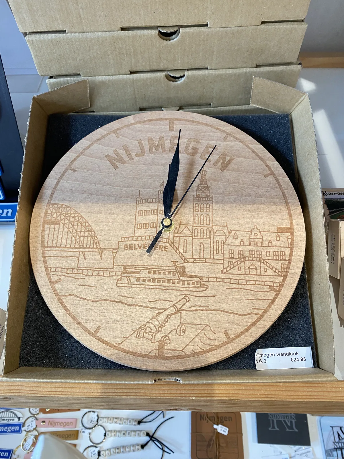 A wooden clock, still in the box. On its face a picture of the city of Nijmegen is engraved. The clock points at one minute past 12.