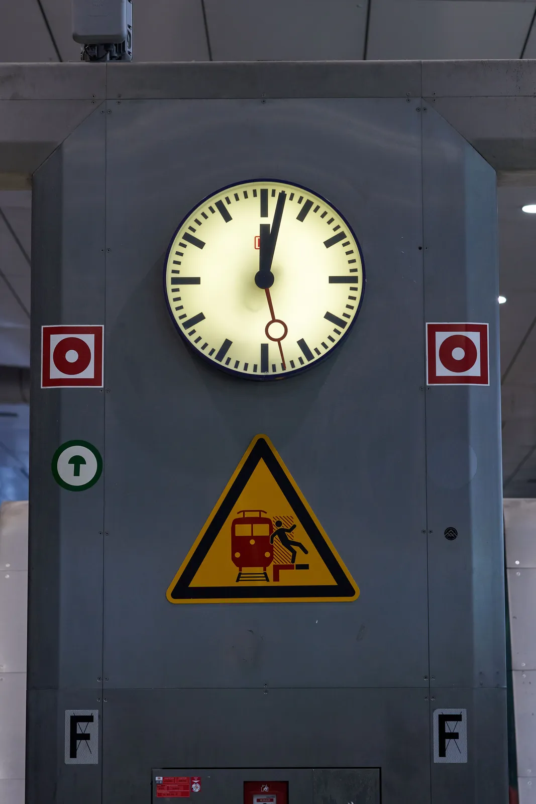 A clock on a metal pillar with stickers on it. According to the clock it is 2 minutes past 12.