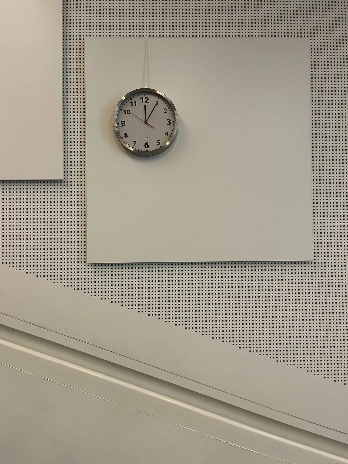 A wall clock is attached to a white square, off center. The white square is attached to a white wall. The upper part of the wall is perforated. A diagonal line separates the perforated part from the non-perforated part. The clock points at 5 minutes past 12.