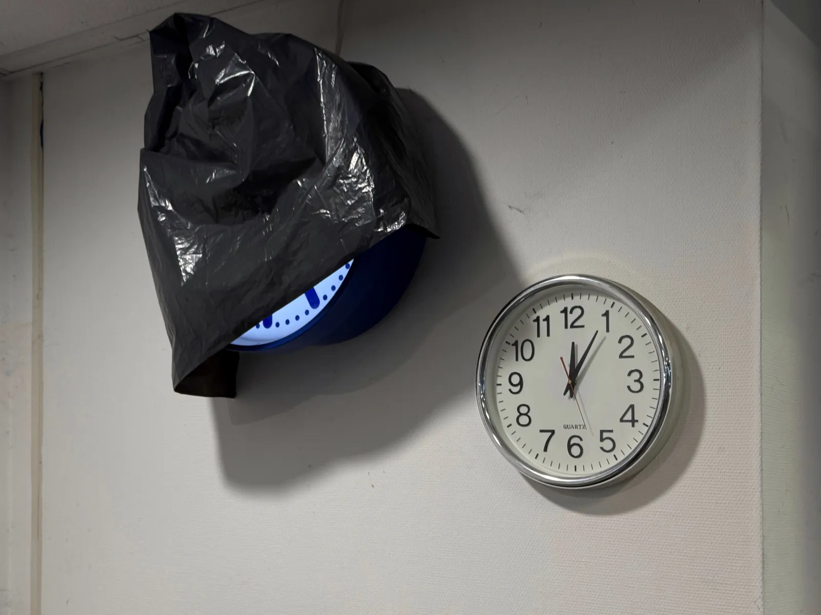 A black plastic bag almost completely covers a lit, modernist station clock. Next to it hangs a simple wall clock which points at 5 past twelve.