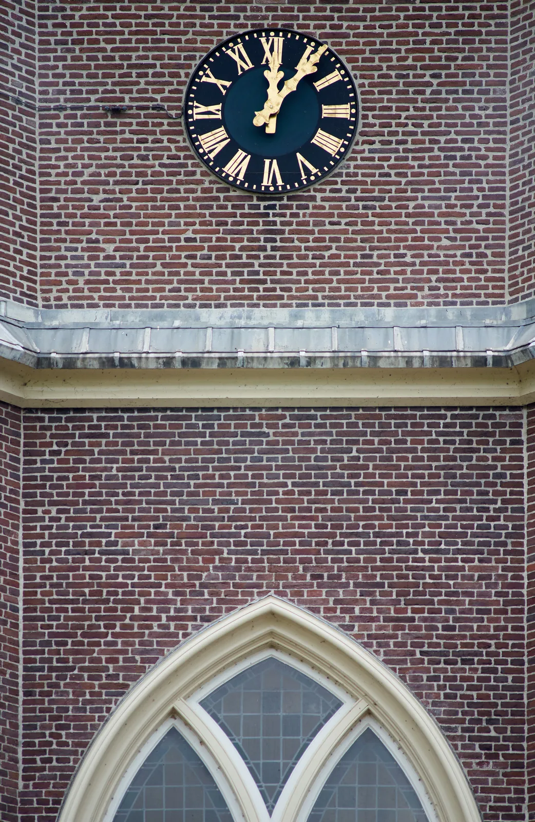 A brick wall with a gothic window and a clock. The clock points at 6 past 12.