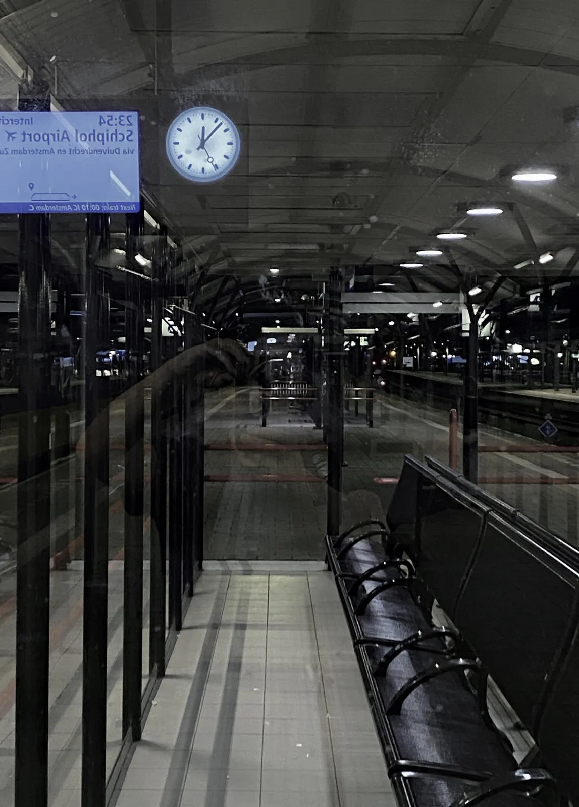On a train station, on a platform, looking at a clock mirrored in the glass of a waiting area. It seems to point at 7 past 12.