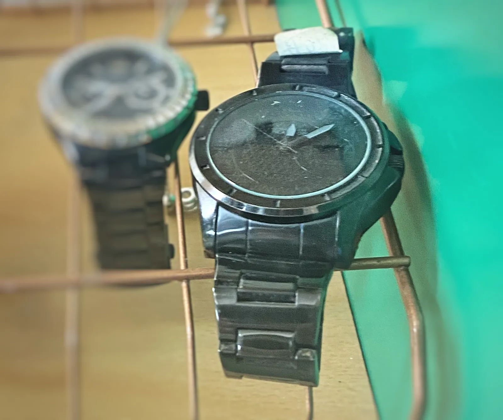 Two wrist watches, large ones. One of the two is in focus. The time on it is approximately 7 minutes past 12.