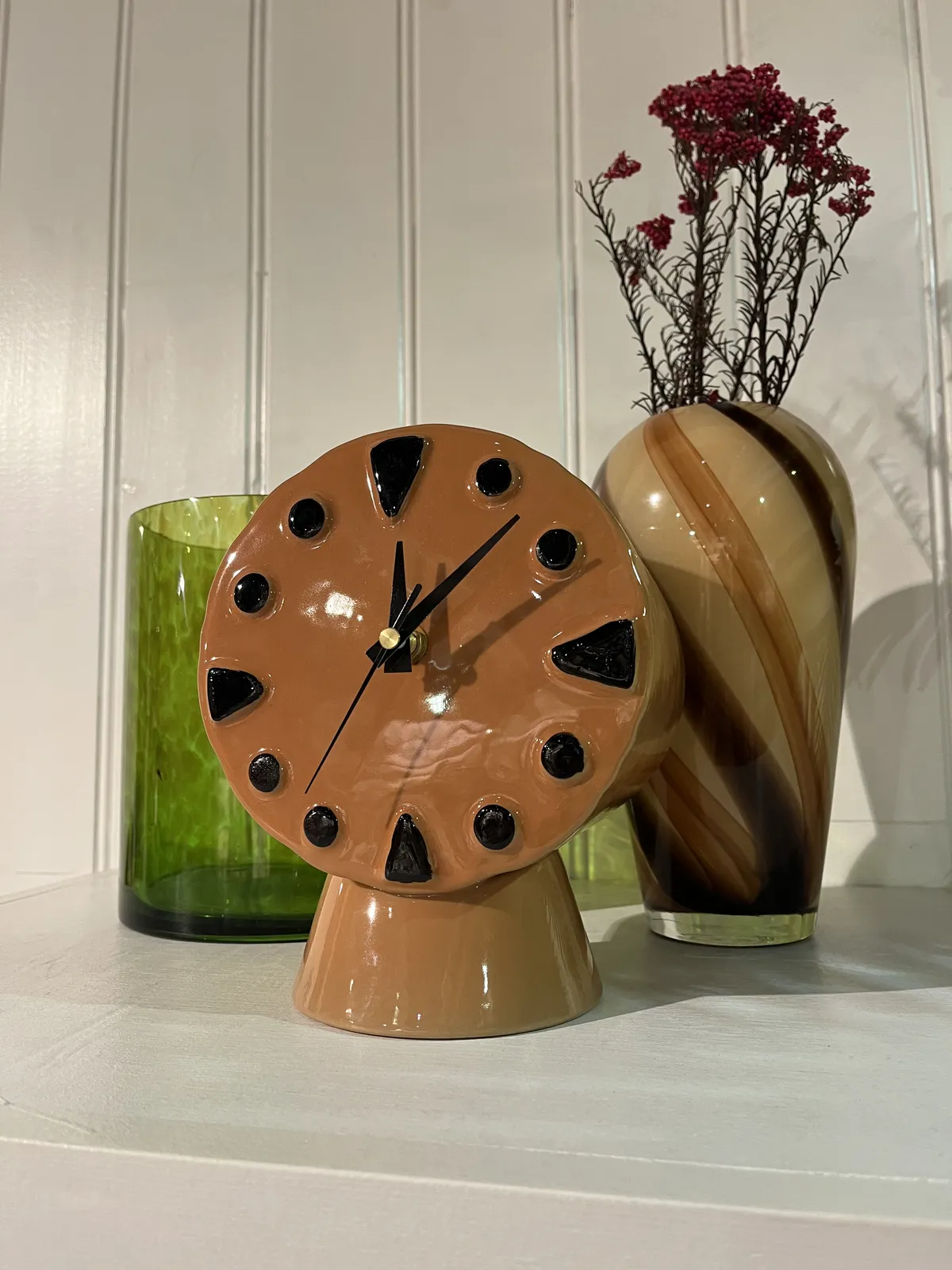 A red ceramic clock in front of a green glass and a vase. The clock points at 7 minutes past twelve.