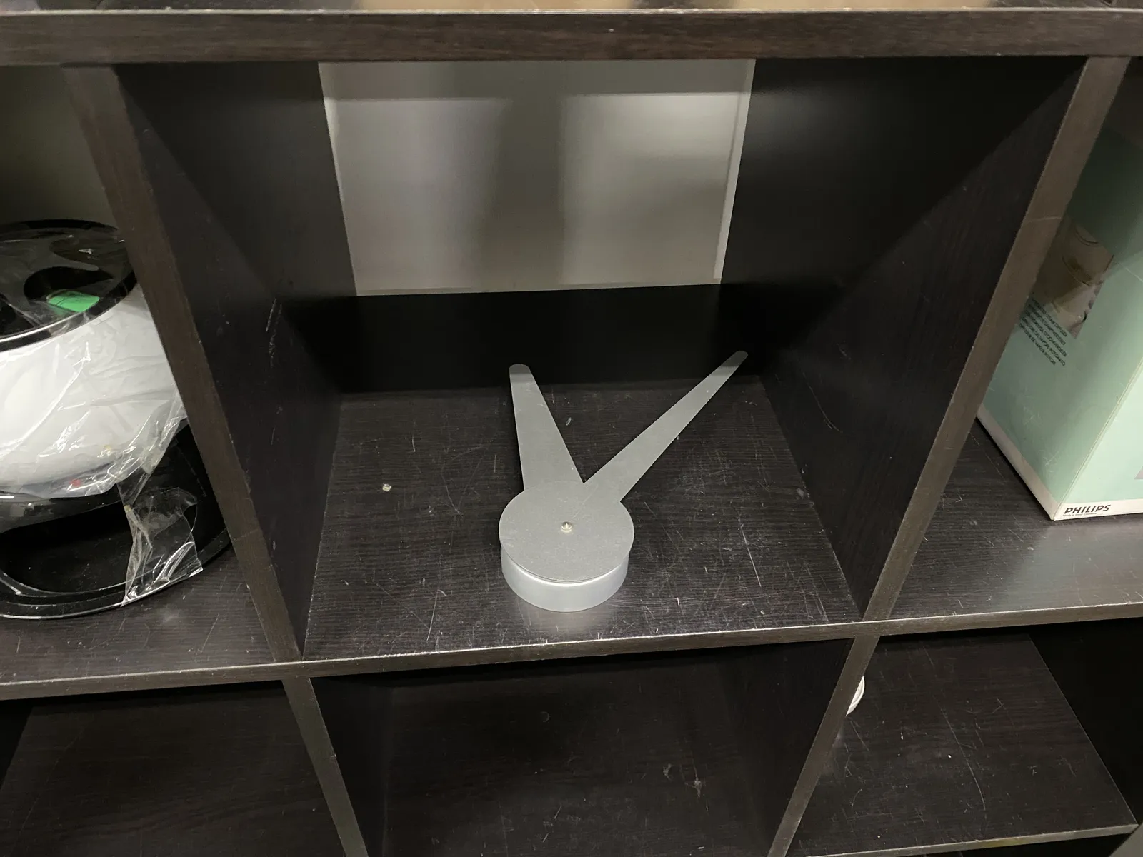A clock that is made of just the two hands. It’s placed in a bookcase. It seems to point at 8 minutes past 12.