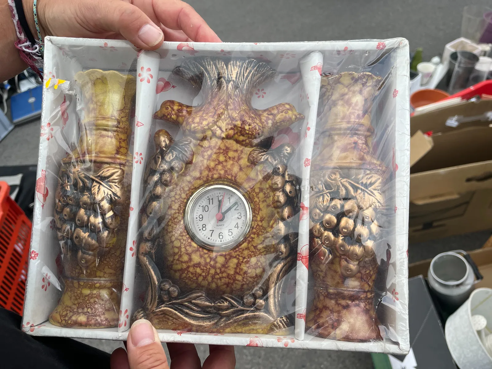 There are two vases, and one clock, still boxed and wrapped in plastic. The vases are placed right and left from the clock in the box. There are birds, leaves, and grapes on the vases and the clock. The grapes and leaves are coloured gold, the rest is a weird mix of orange and red, in a vein-like pattern. The clock looks a bit like a pineapple and it points at 8 minutes past 12.