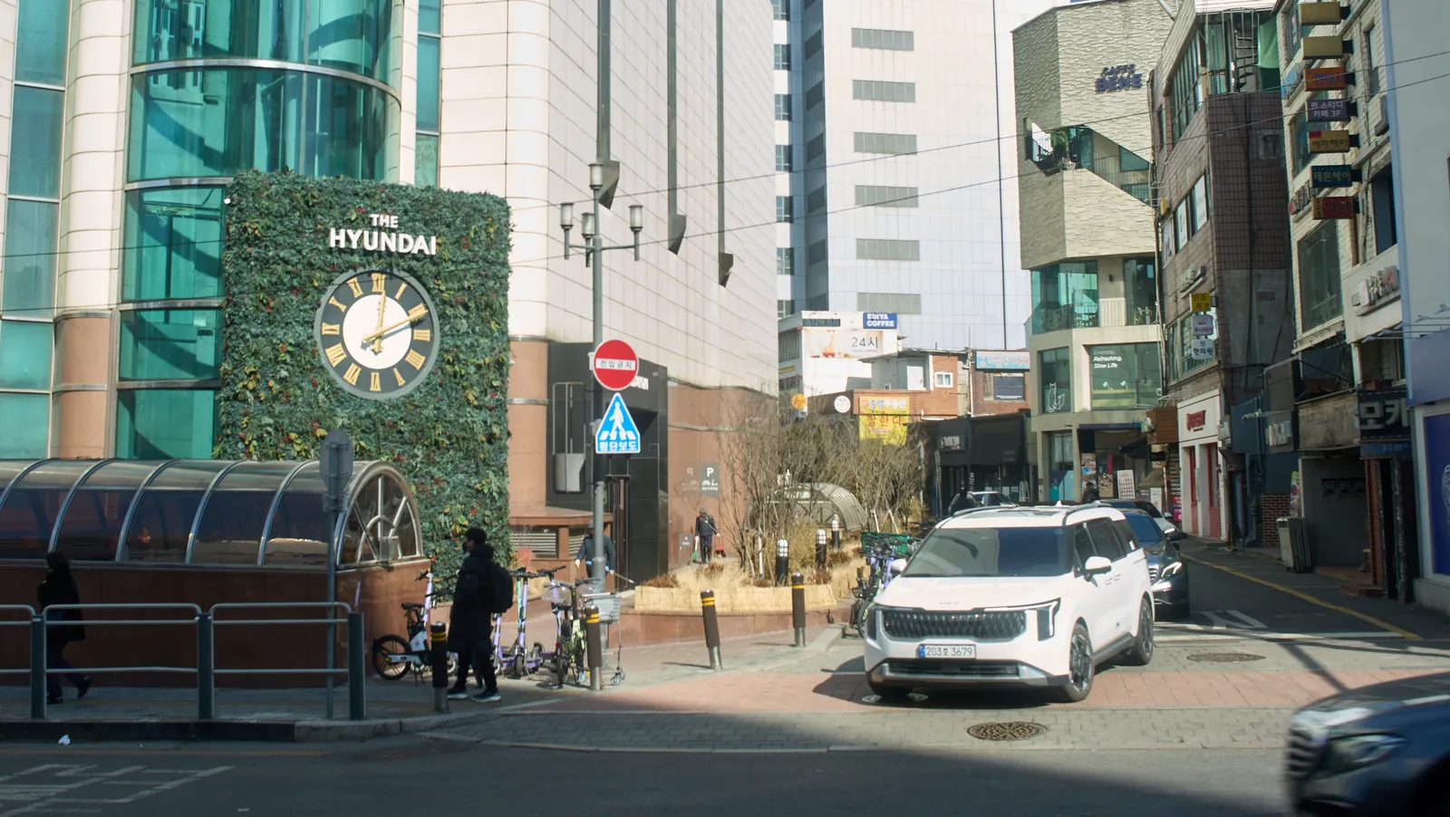 A street with lower buildings on the right, and a very high building on the left. In front of the building, in a hedge, there’s a rather large clock. It points at 11 minutes past 12