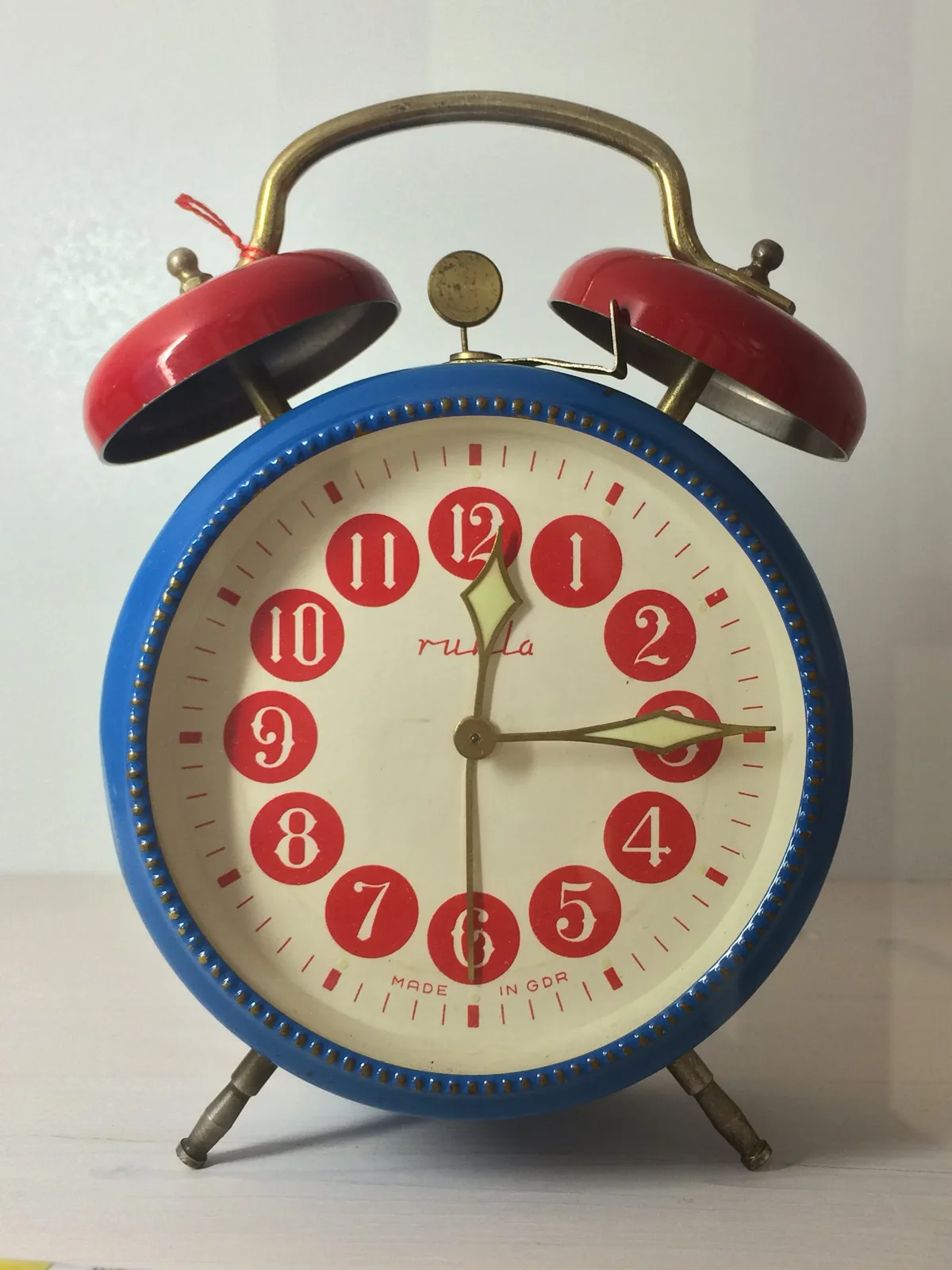 A mechanical, old fashioned alarm clock, the one with bells on top. It’s colourful, blue, white and red, with ornamental and various typography. It’s made in GDR, it says on its face. It points at 14, almost 15 minutes past 12.