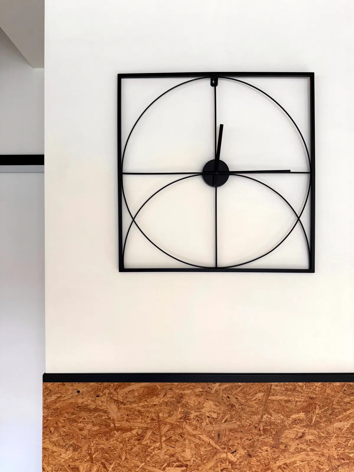 On a white wall there’s a black square. In this square there’s a circle touching all sides, and half a circle at the bottom, all black as well. There’s also a black cross that divides the square into four smaller squares, all the same size. This object is in fact a clock. The two black hands point at 14 minutes past twelve.