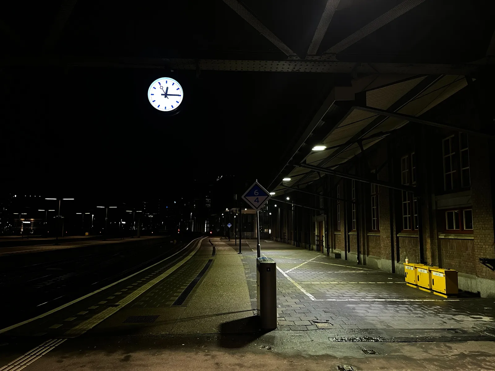 An empty, deserted train station at night, it is very dark. A station clock is lit. It points at a quarter past midnight.