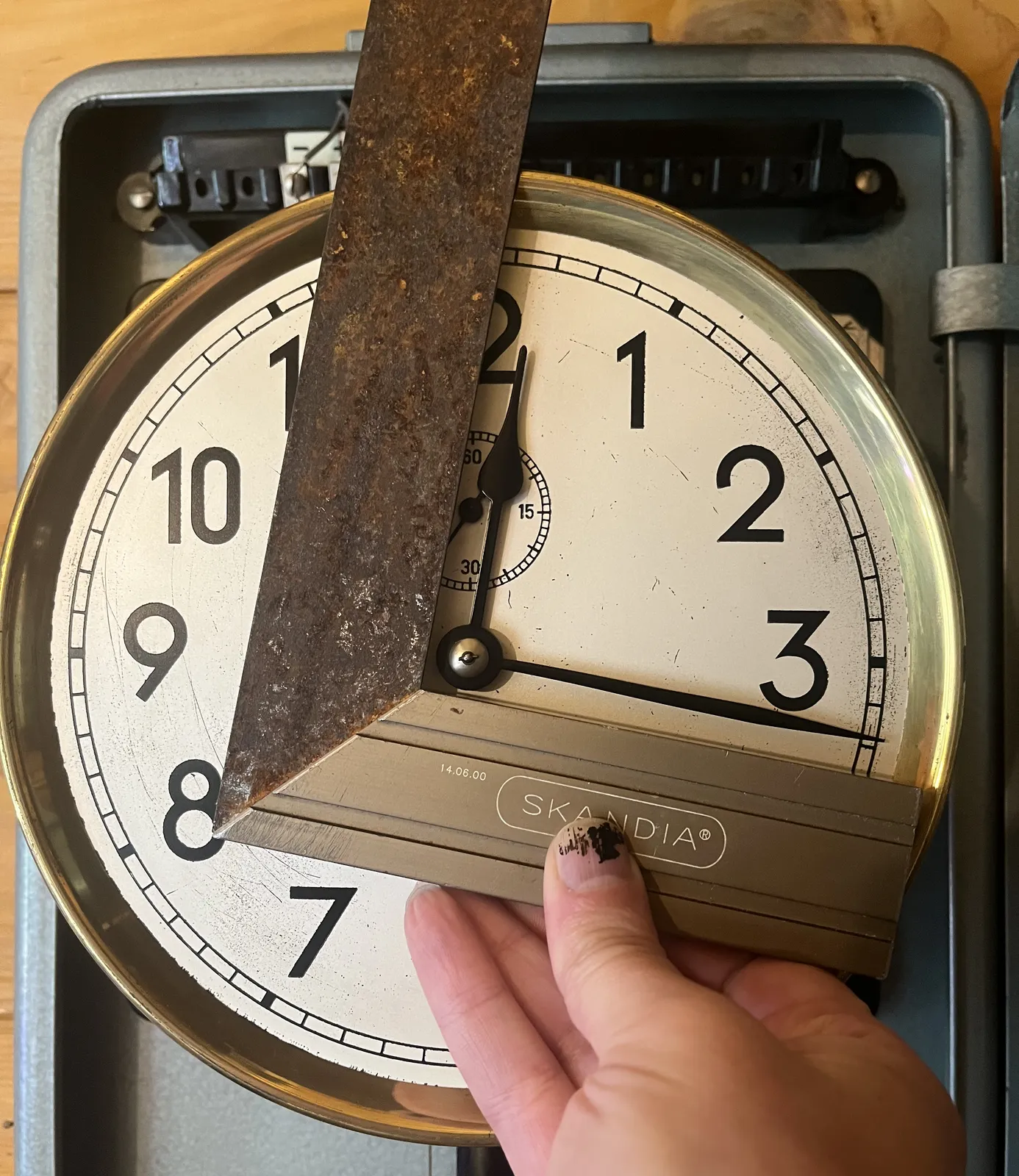 Someone is holding a try square in front of a clock, to show that the two hands of this clock form a perfect 90 degree angle at a little over 16 minutes past 12