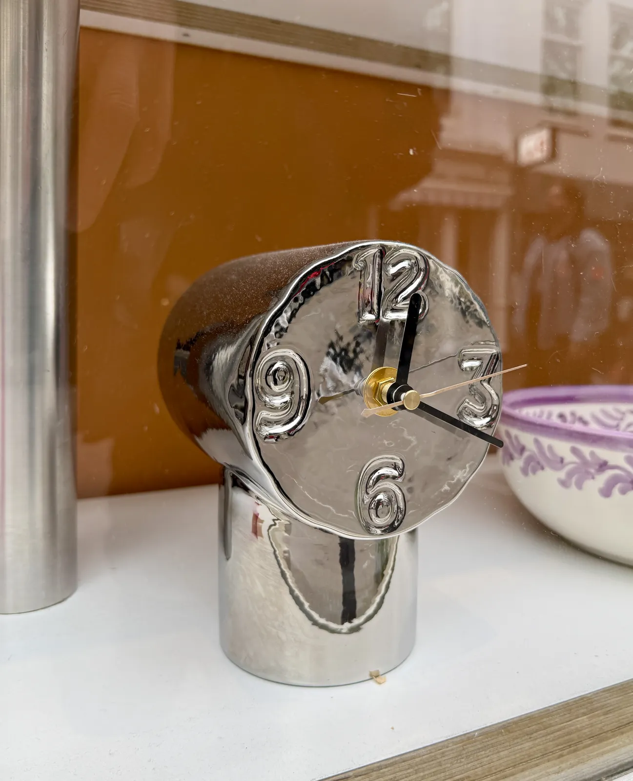 A shiny clock made from two cylinders. The numbers 12, 3, 6 and nine are embossed in it. It points at eighteen minutes past twelve.