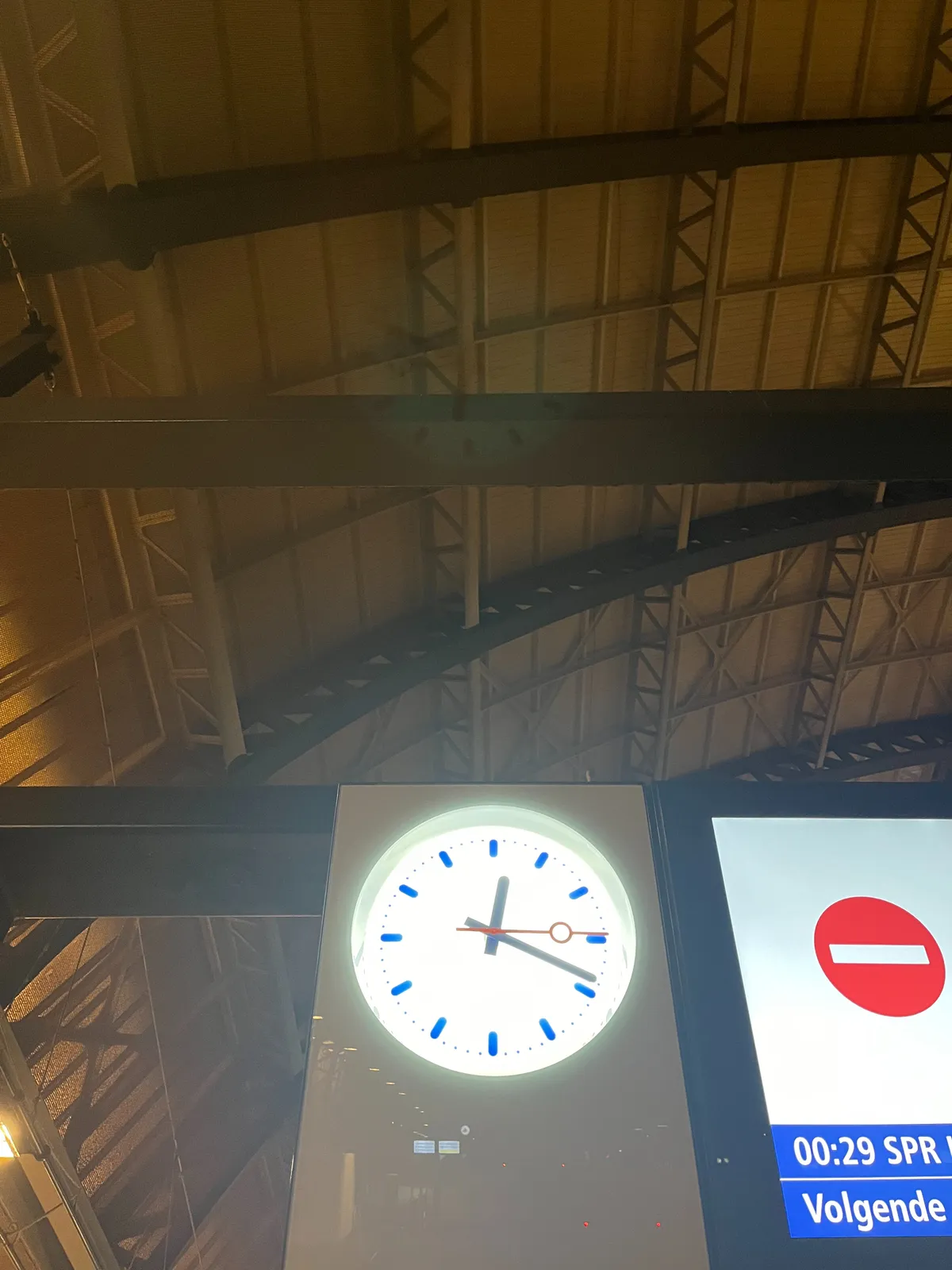 Looking up, in the background you can see a part of the cast iron dome of the Amsterdam Central train station. In the foreground there’s a part of a train display with a street sign on it that indicates not to enter this train. Next to the display there’s a clock. It points at 19 minutes past 12.