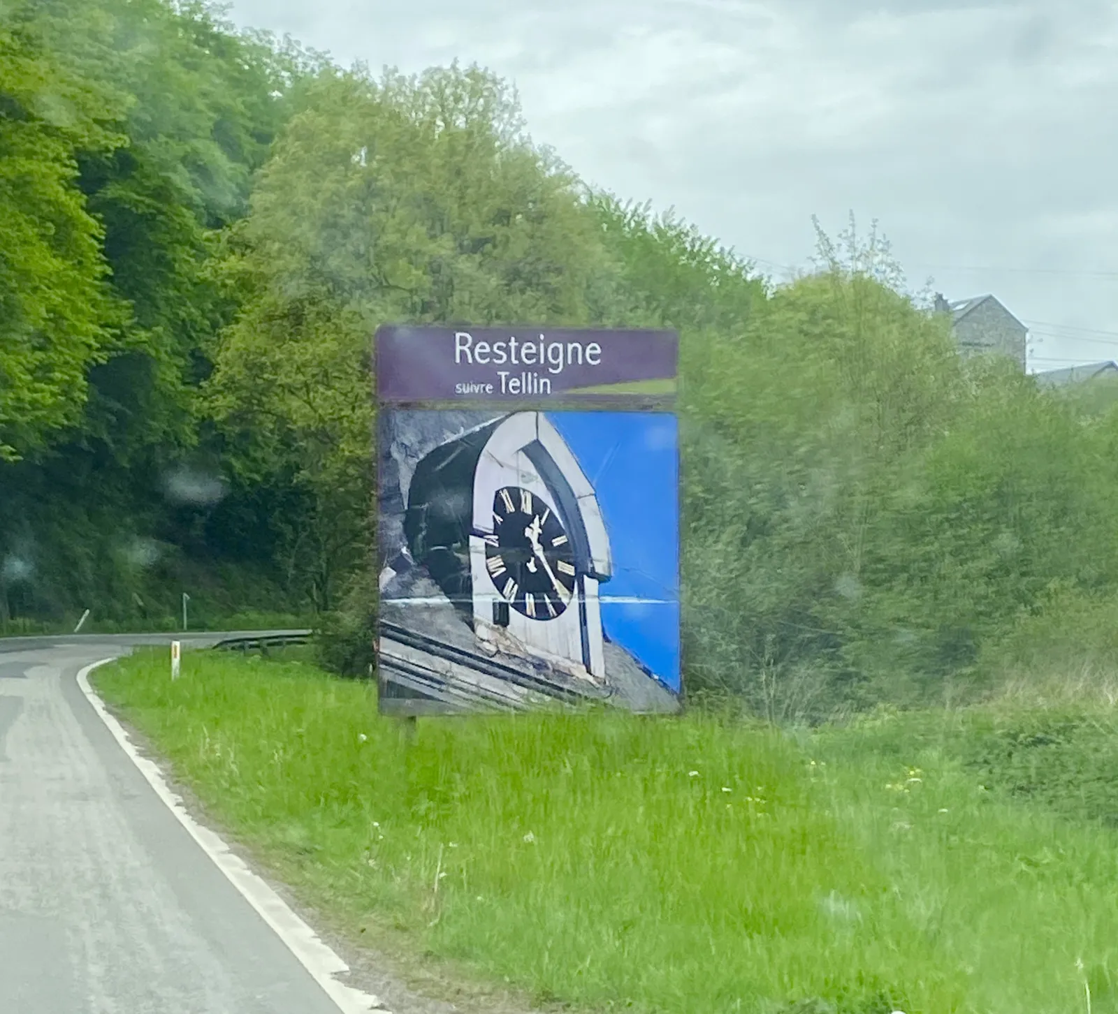 A large road sign. It shows a close-up of a clock on a building. This clock can be found in Resteigne, when you take the exit towards Tellin. The clock on the billboard points at 21 minutes past 12.