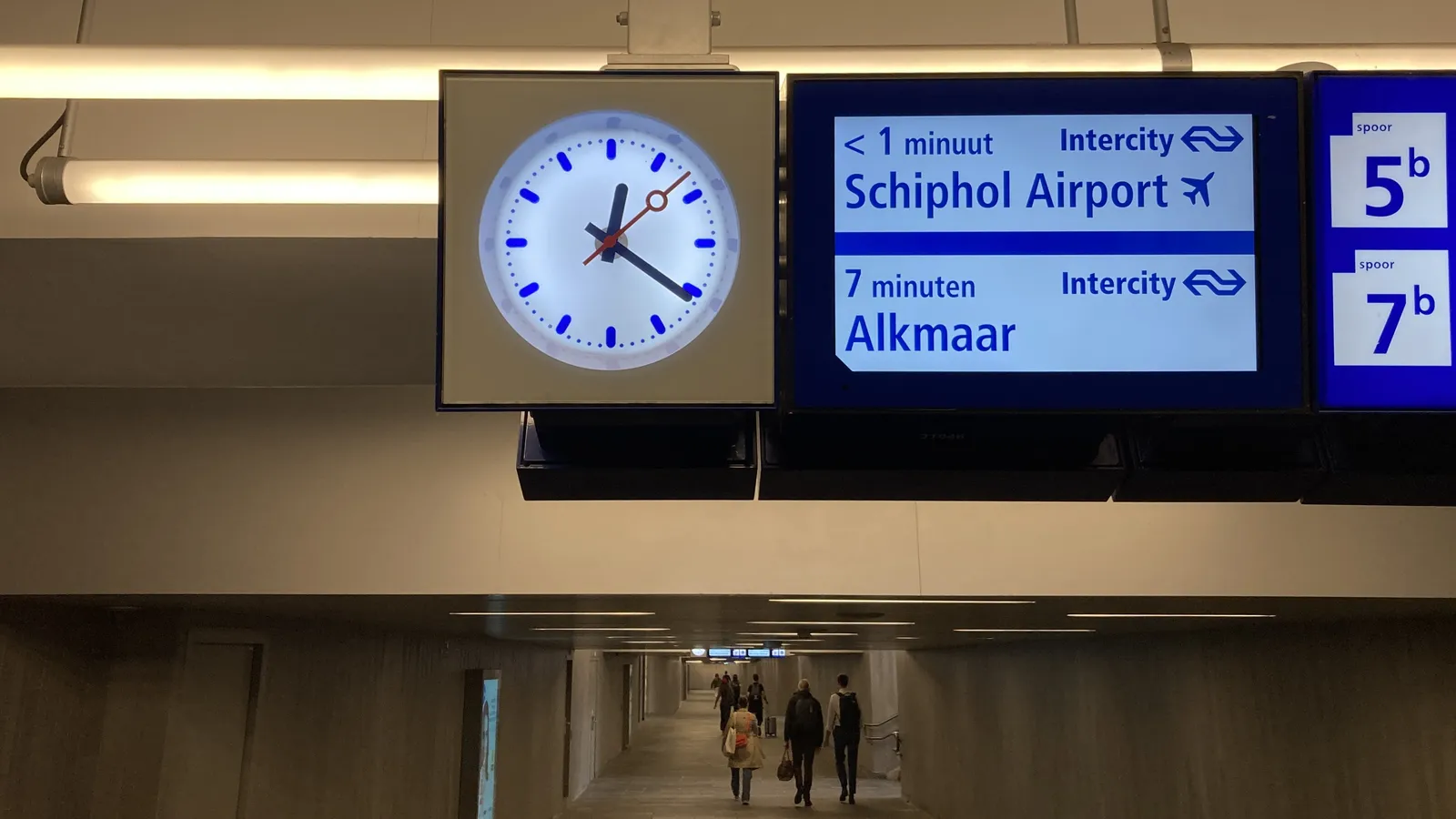At the bottom of the picture there’s a peek into a pedestrian tunnel. Most of the picture is taken by a display that shows that two trains will be leaving soon. Next to this display there’s a clock that shows it is 21 minutes past 12.