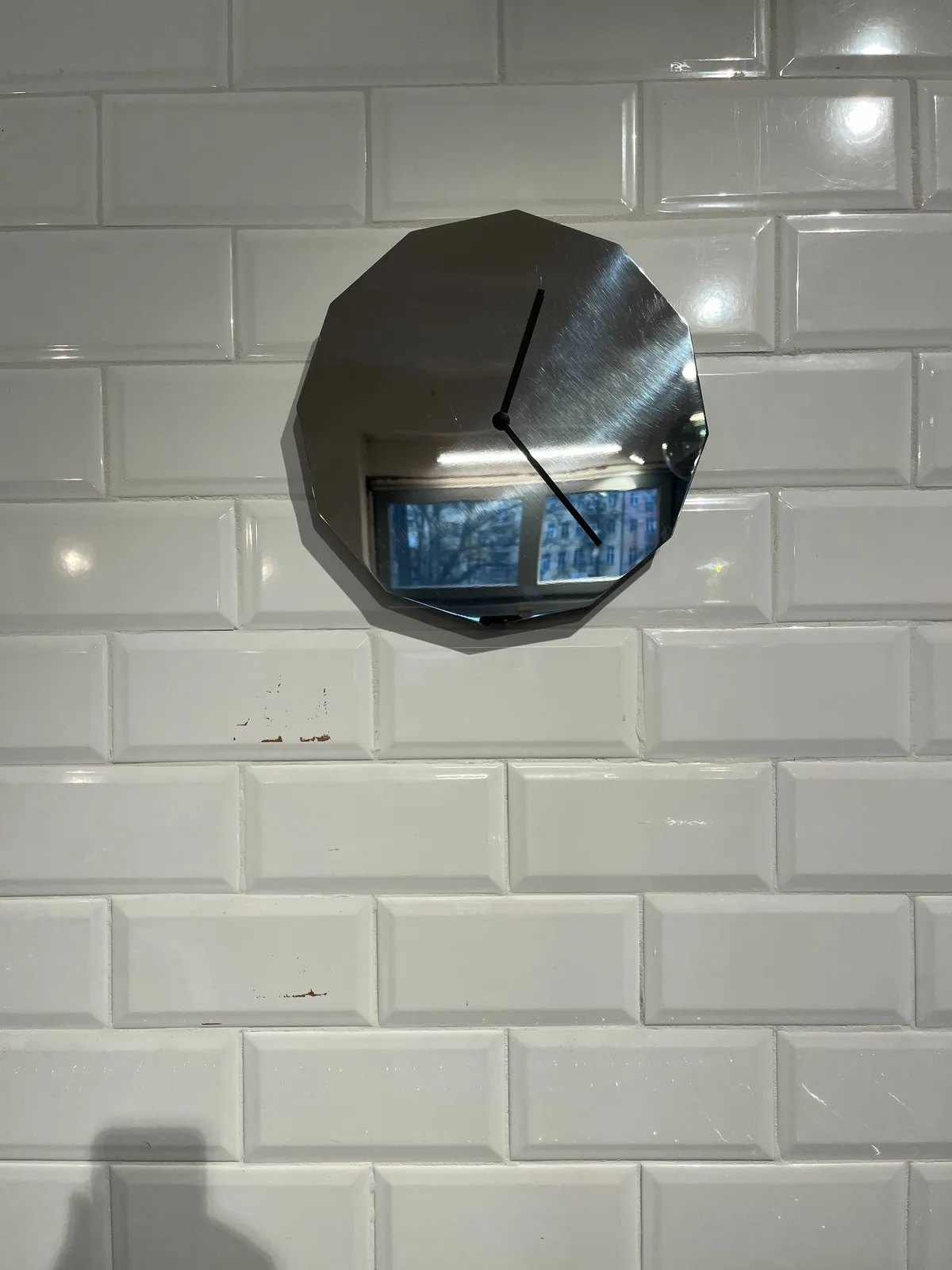 On a wall made from white tiles, there’s a clock made from a mirror with twelve corners. The black hands point at approximately 22 minutes past 12.