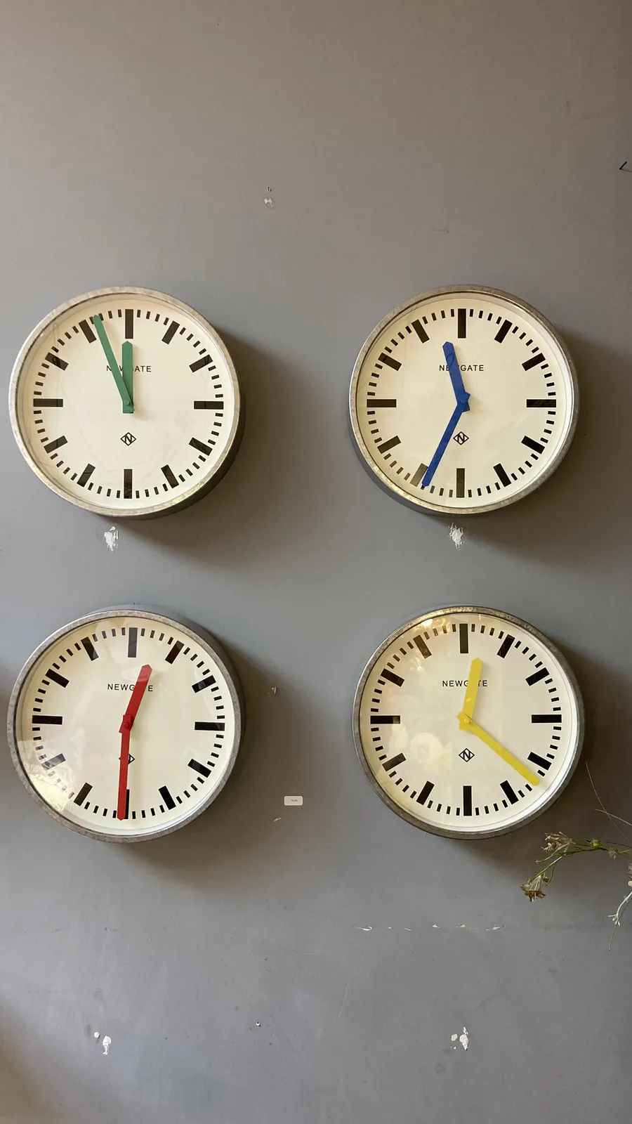 Four almost identical modernist clocks on a gray wall. The top left one has green hands, and it points at four minutes to twelve. The top right one has blue hands, and it points at 34 minutes past 11. The one on the bottom left has red hands, and it points at half past 12. And finally, the one on the right bottom has yellow hands, and it points at 22 minutes past 12. 