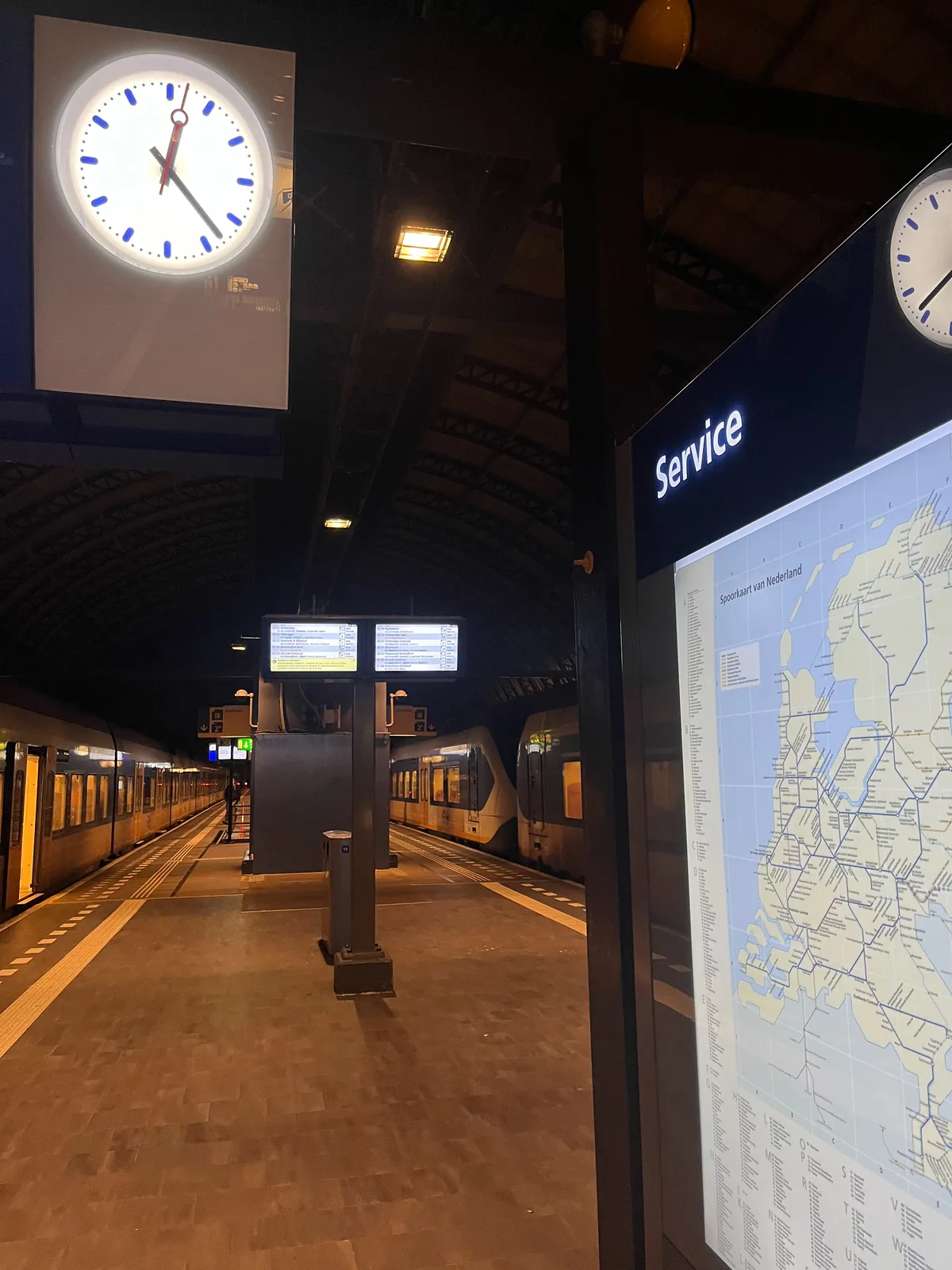 On the right threre’s a large display with the map of the Netherlands and its railway system on it. The word Service is printed over it. You can also see a reflection of a clock on this display. In the background two parked trains and an empty platform. There’s also a clock which points at 23 minutes past 12.