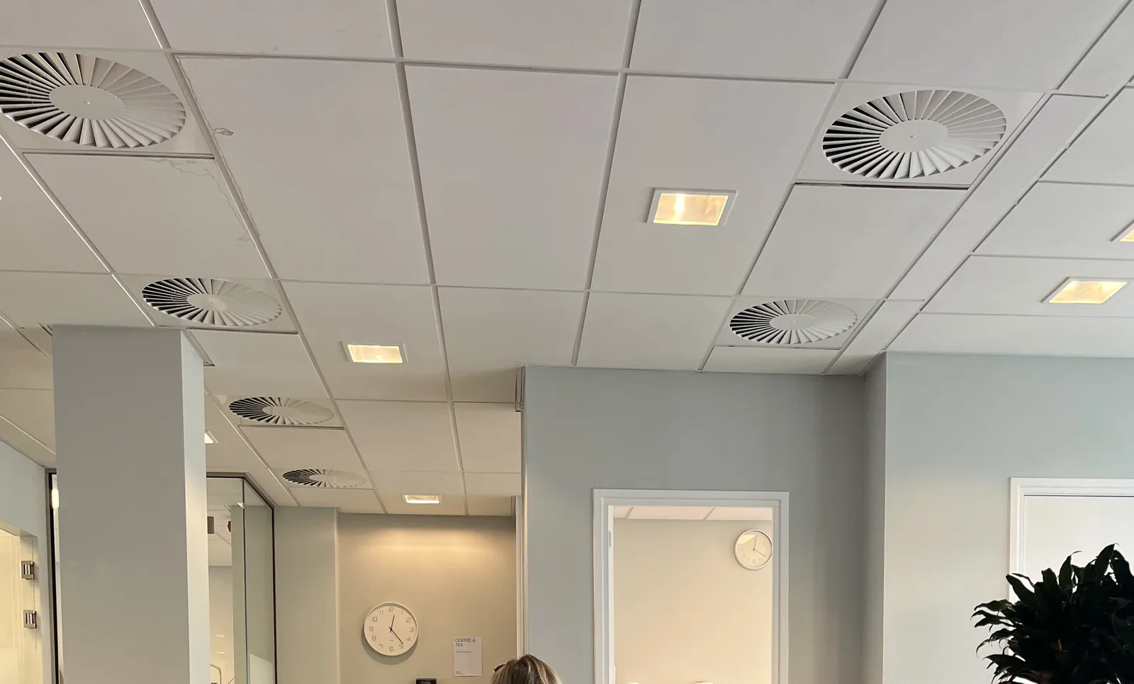 Looking at a white, dropped ceiling. There are ventilator like holes in it. In the back of this room you can peek into two other rooms, both with a clock on the wall. The clock on the left points at 23 minutes past 12, the one on the right at 20 past.