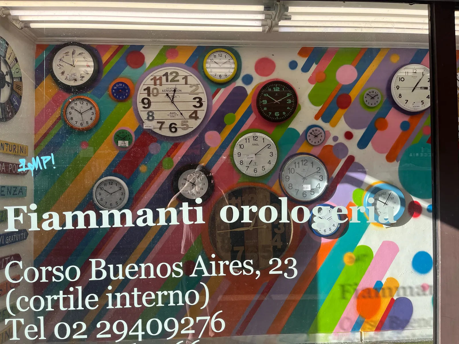 The very, very colourful shopping window of Fiamanti Orologeria on the Corso Buenos Aires 23. It says so in large white letters in the glass. There are many, many clocks on display, all very colourful. One of the clocks points at 24 minutes past 12. Another clock points at 16 minutes to one. A clock points at 6 minutes past one. One of the clocks points at 11 minutes to two. Another one points at 9 minutes to two. Then there’s a clock that points at 8 minutes past 7. There’s also a clock that points at 9 past 8. And there’s a clock that points at 5 past 10. Also, there are clocks that point at 9, 10 and 11 minutes past 10. And finally there’s this one clock that points at 11 minutes to 12.