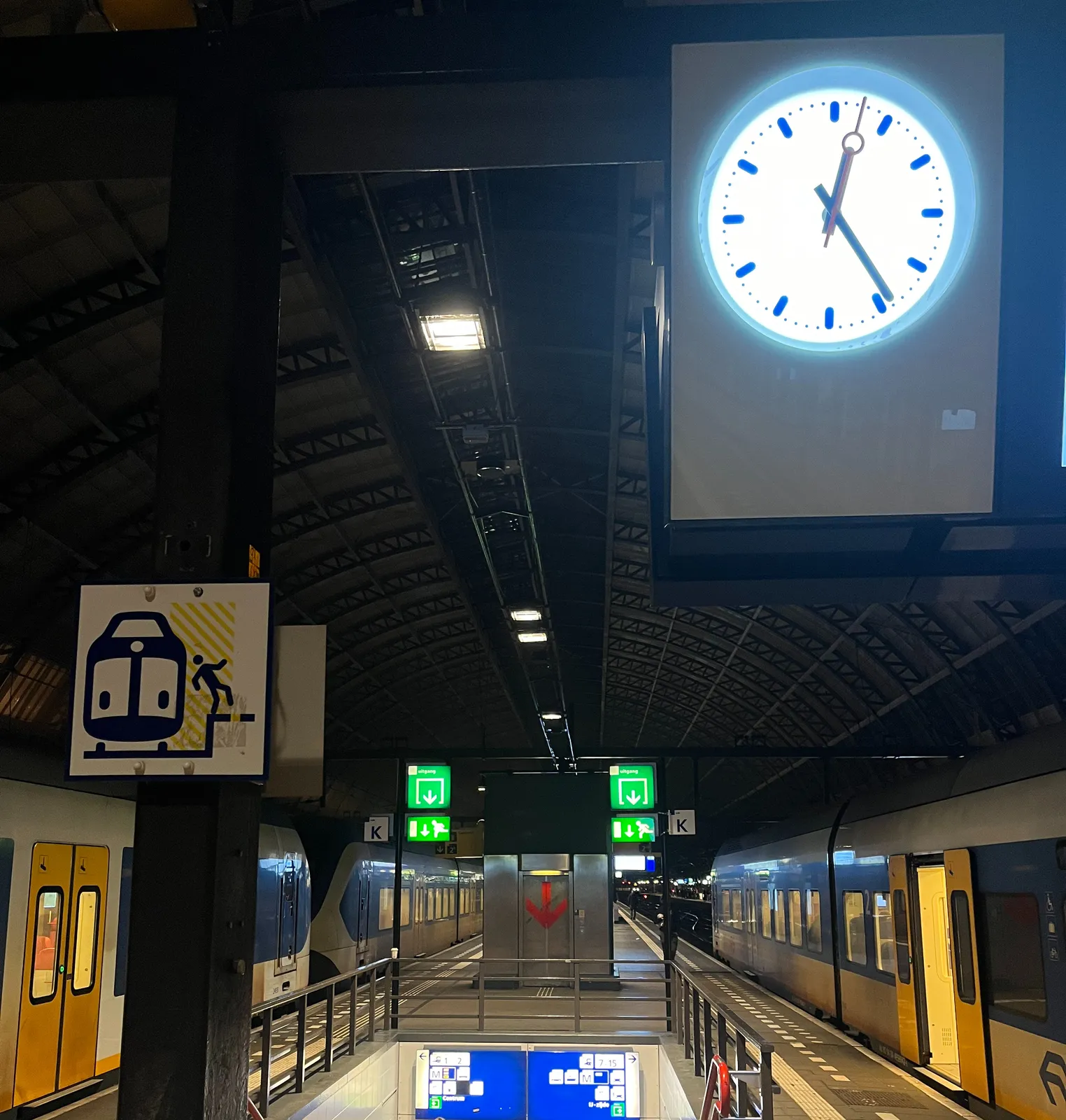 A sign that shows a train and a person falling of a platform. Parked trains on an empty platform, with stairs going down. In the background a huge red arrow pointing down. And there’s a clock that shows the time as 24 minutes past 12.