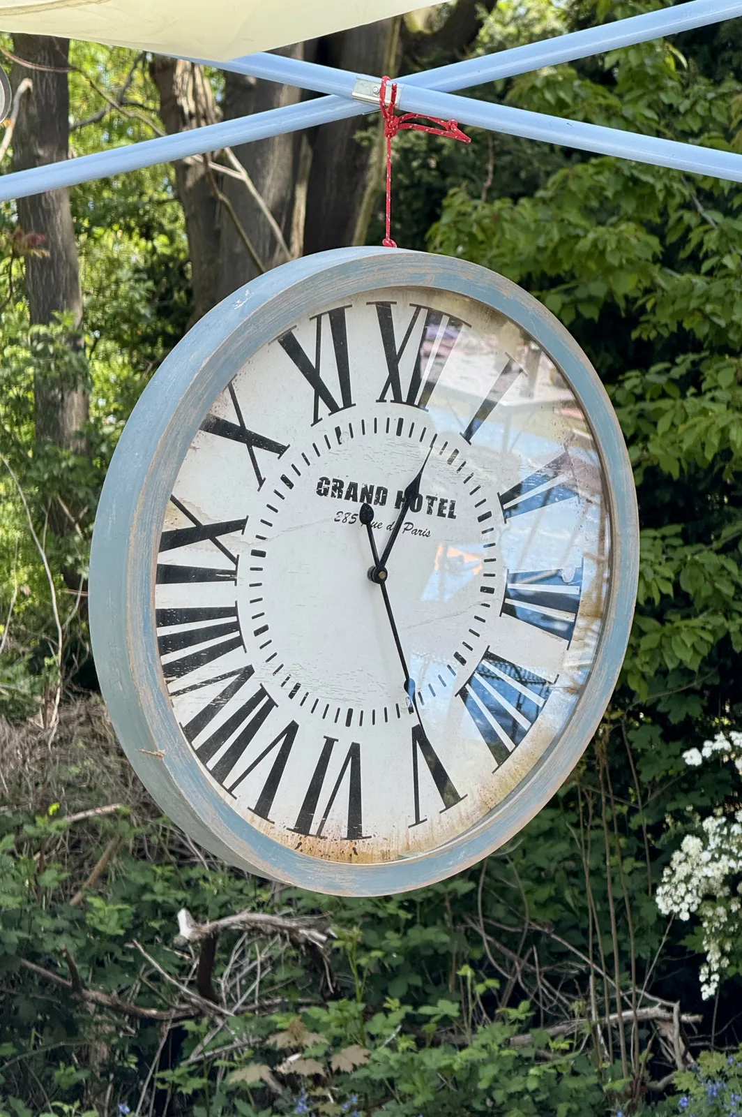 A large wall clock with large roman numerals hangs in mid air from a red shoo lace. In the background there’s a forest. The clock points at 24 minutes past twelve.