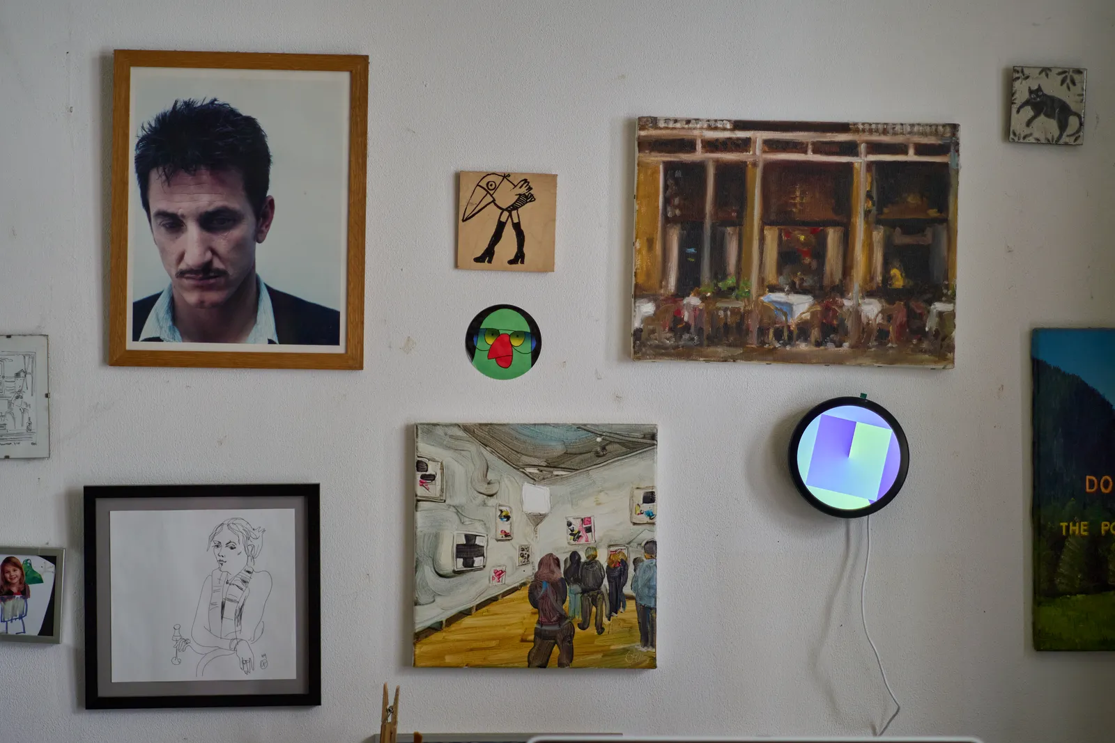 A white wall with all kinds of works of art on it, amongst others a picture of a man with a moustache and a round display with an abstract image on it, made from two gradients on top of each other. If you look carefully at this round display you might see that it is in fact a clock, and the gradients form the hands, which point at 25 past twelve.