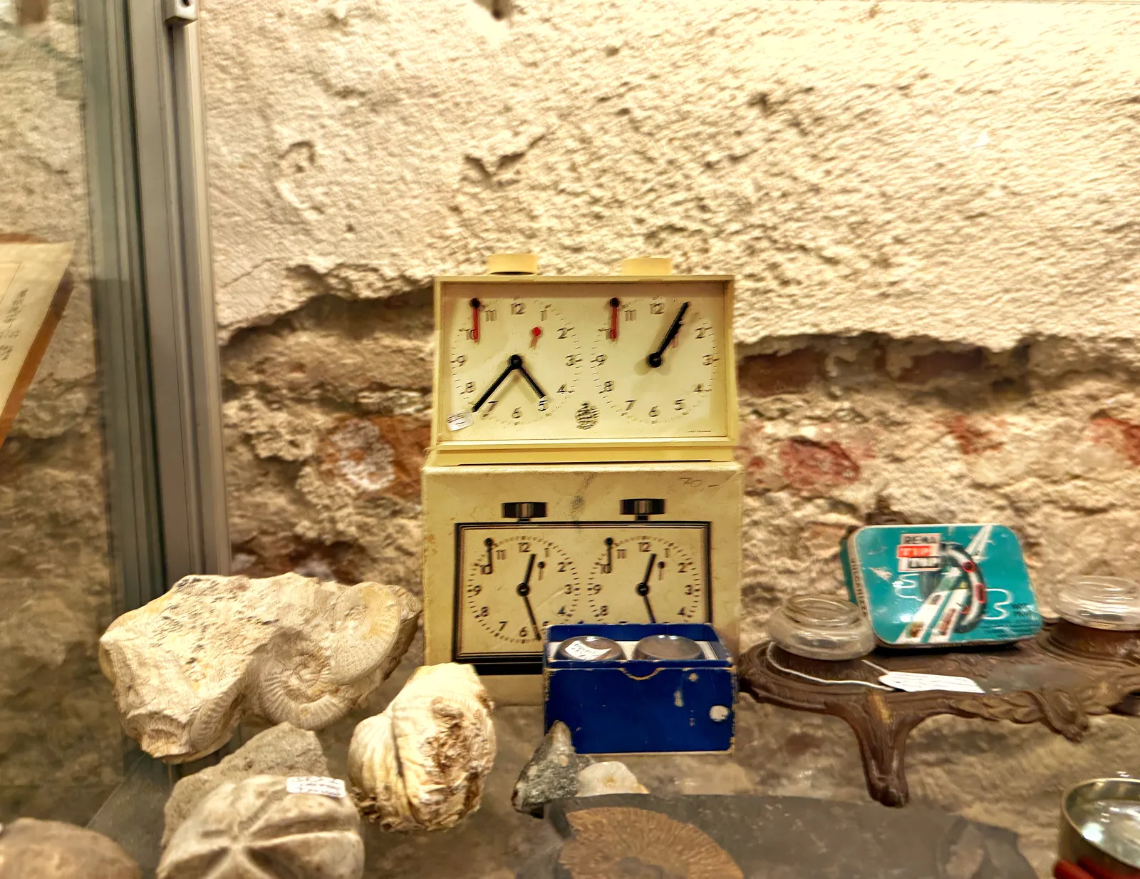 On top of the box with a chess clock printed on it, stands this same chess clock. The clocks on the box both point at 27 minutes past twelve. The left clock on the real chess clock points at 37 minutes past four, and the one on the right at four minutes past one. Around this clock and the box there are fossils and other old things.