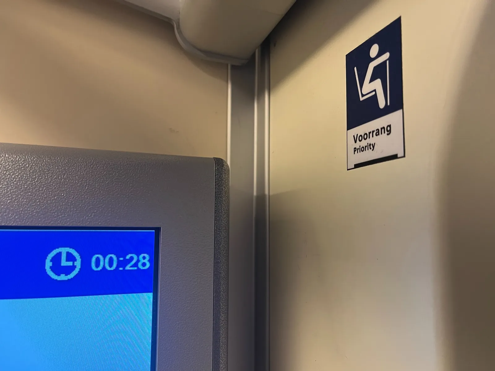 Looking at a corner. On the right wall there’s a sticker of a person with a walking stick, sitting. Below it the words voorrang and priority are printed.  On the left side there’s a small part of an information display that shows the time. It is 00:28.