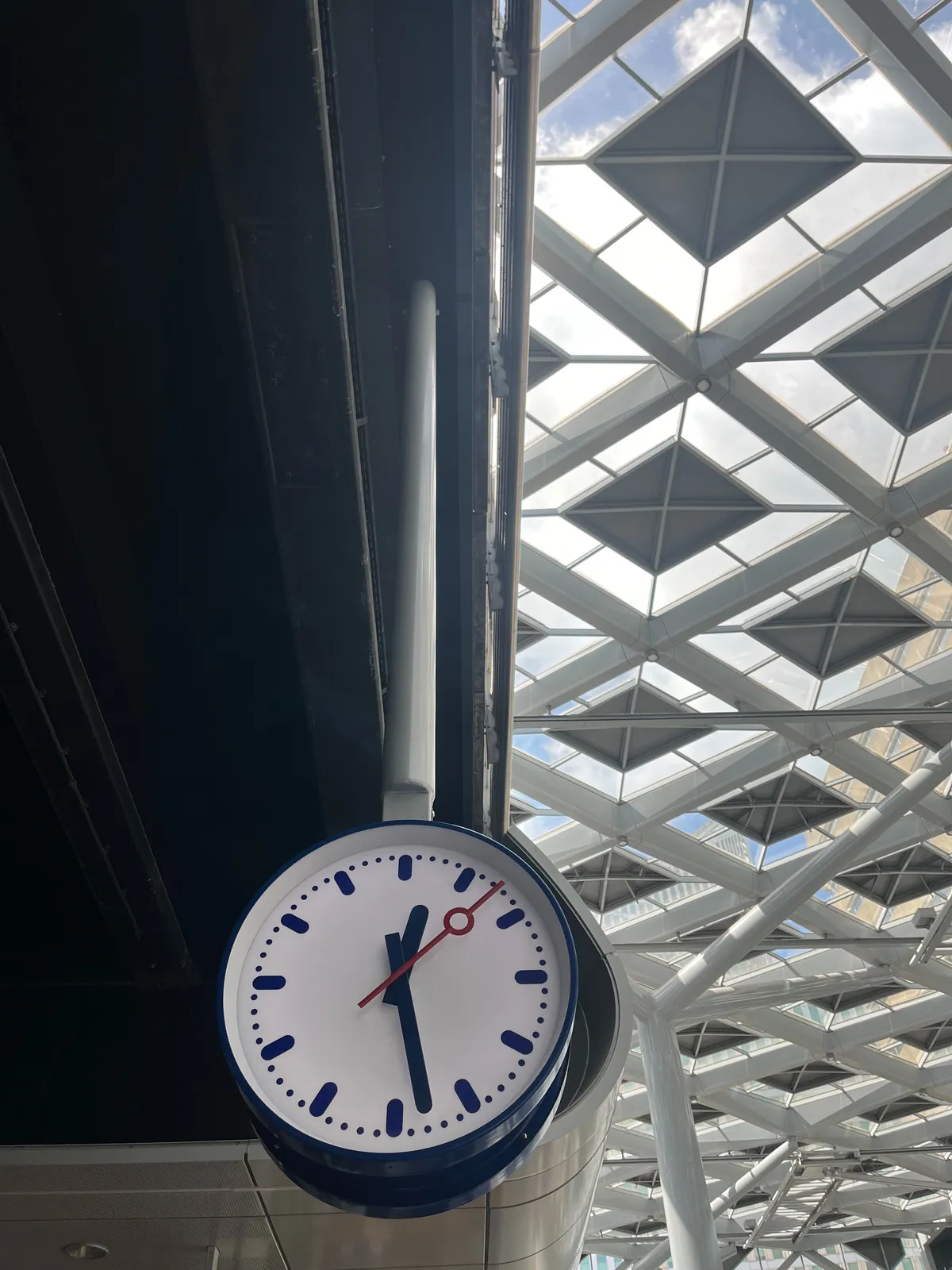 A station clock hanging from a ceiling. It points at 28 minutes past 12.