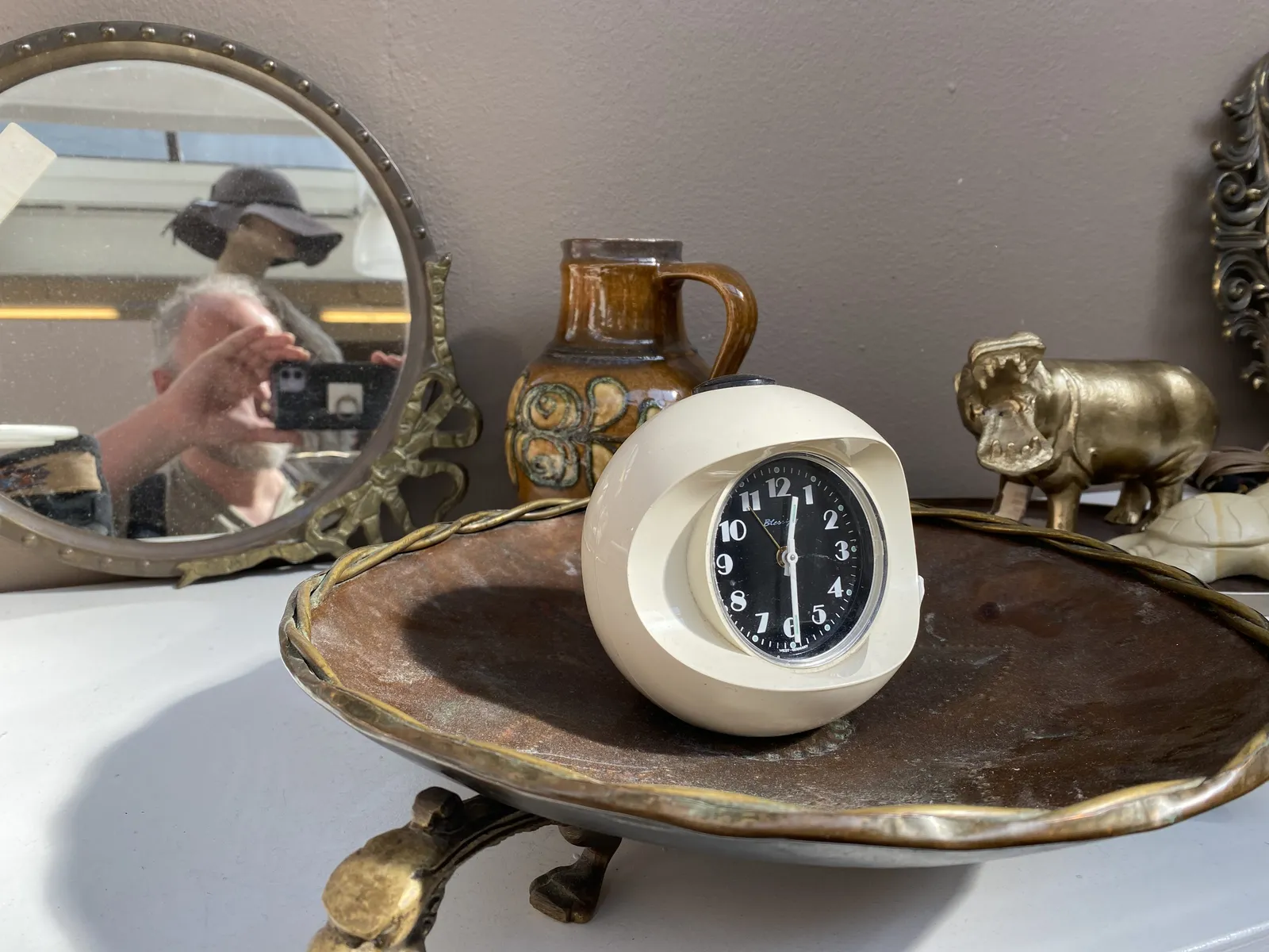 There’s a little statue of a hippopotamus, there’s a little vase and there’s a mirror. In the mirror you can see the photographer taking the picture. There’s also a dish with a plastic space age clock on it. The clock points at 29 minutes past 12.