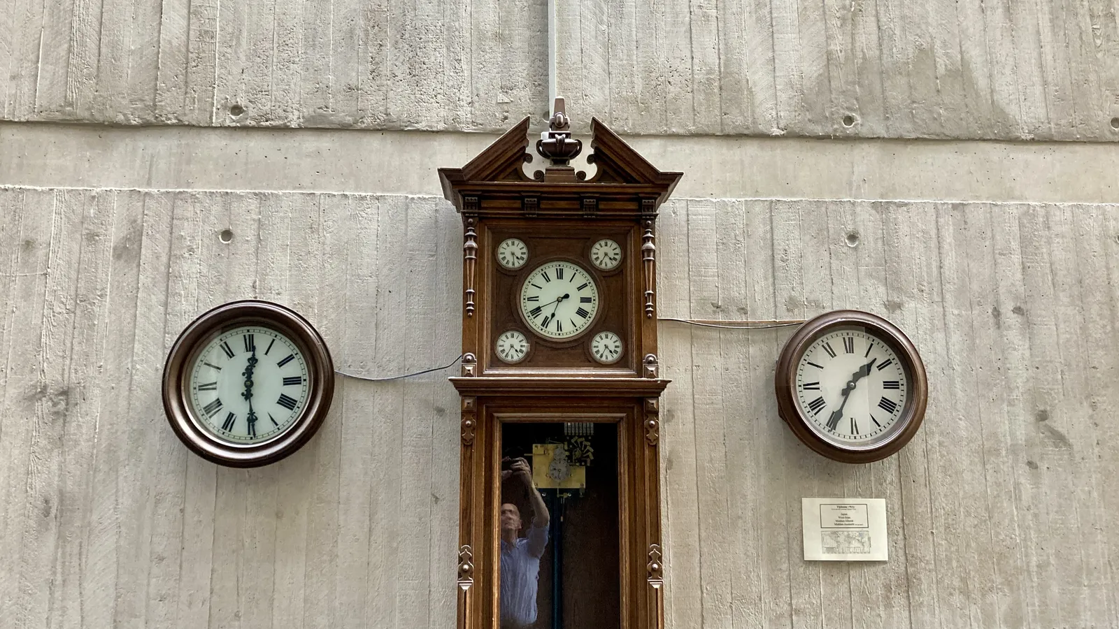 A work of art. It consists of multiple clocks. In the center there’s an old, wooden, standing clock which actually has five clocks. The main clock points at 41 minutes past 6. Around this main clock there are four small clocks. Three of them point at 35 past 4. One of them lags a little behind: it points at 29 minutes past 4. On the left of the standing clock there’s a wooden wall clock that points at 29 minutes past 12 and on the right there’s a similar clock that points at 34 minutes past one. The photographer can be seen in the reflection, in the glass of the large standing clock.