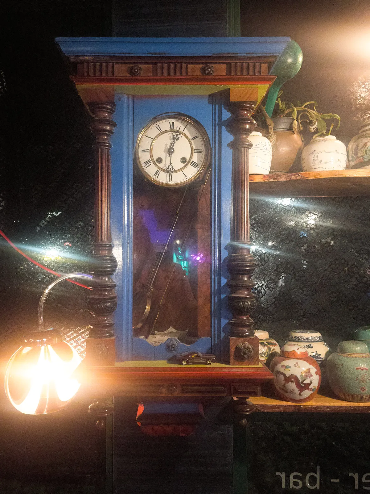 Surrounded by lights, and some nice little ceramic pots, ther’s a blue, wooden, mechanical wall clock, with some nice ornaments. It points at half past 12.