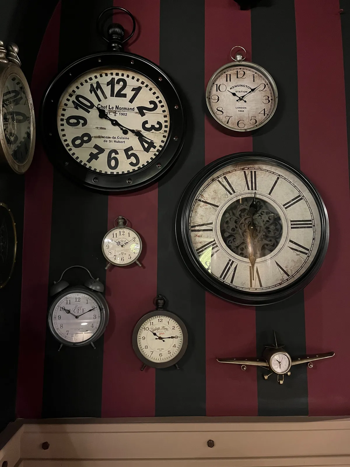Quite a few clocks on a wall. The wall has vertical, wide stripes, black and dark red. One of the clocks points at half past 12. Then there are a few that point at 9 or 10 minutes past 10. There’s a clock that points at a quarter past 10 and there’s one that points at 18 minutes past 10. And finally there’s a smaller clock, shaped like an airplane, that points at 31 minutes past 10.