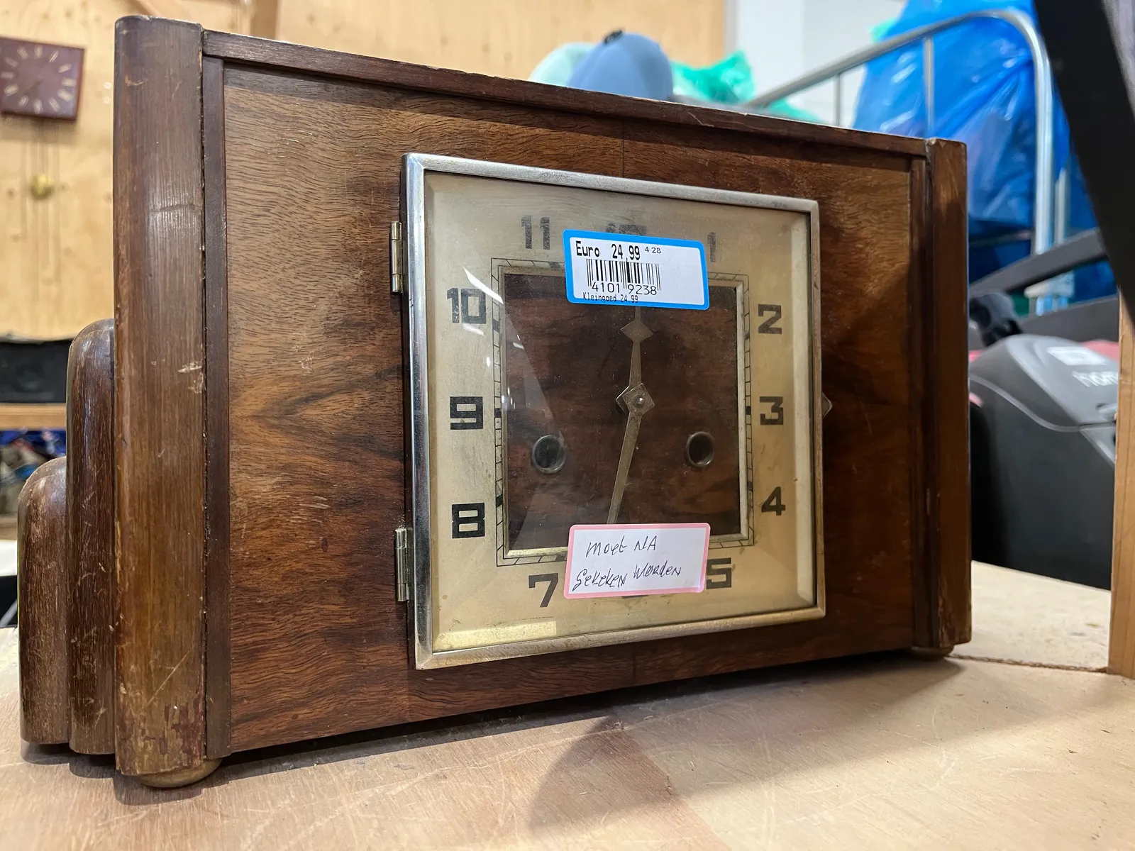 A wooden, art deco mantel clock for sale for 24,99 euros. It needs some repairs, another sticker says. It points at 32 minutes after twelve.