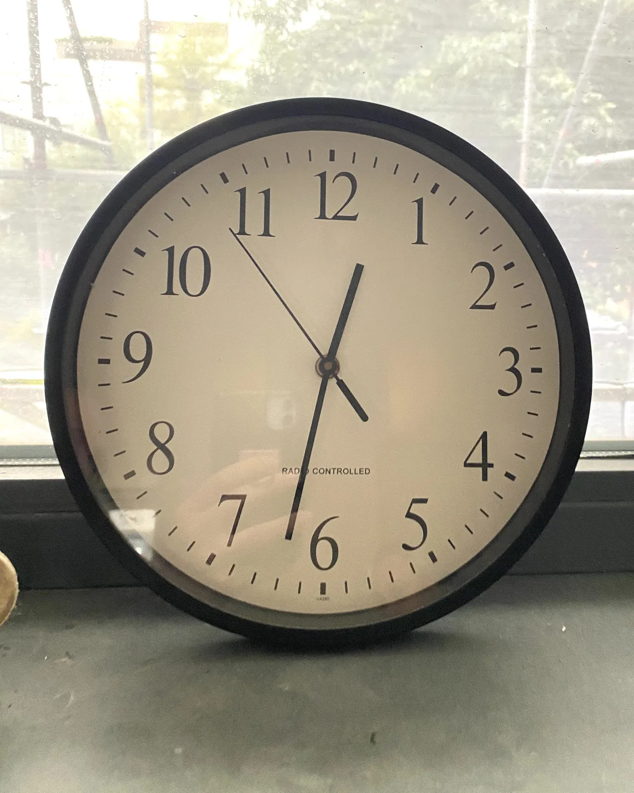 A clock, it says radio controlled on its face, in front of a window. On the clock it is 28 minutes to one.