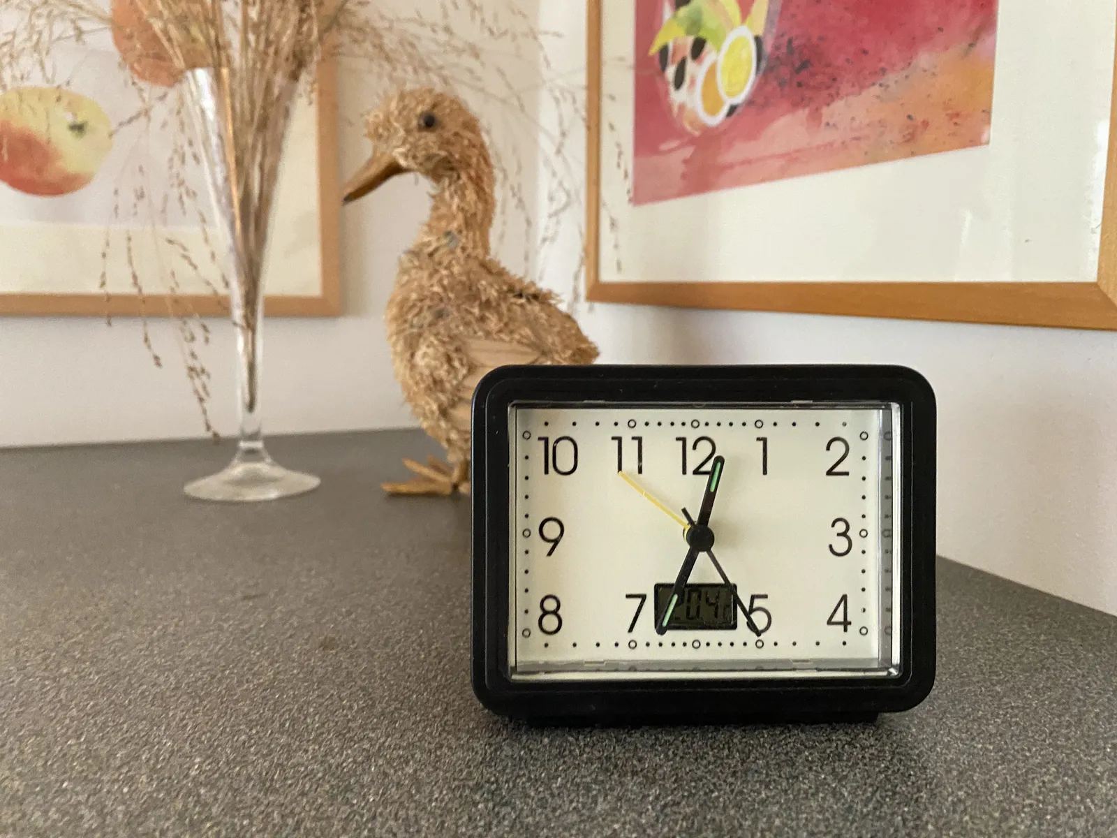 A clock on a desk. In the background there are some paintings and a duck made from straw. The clock points at 27 minutes to one.