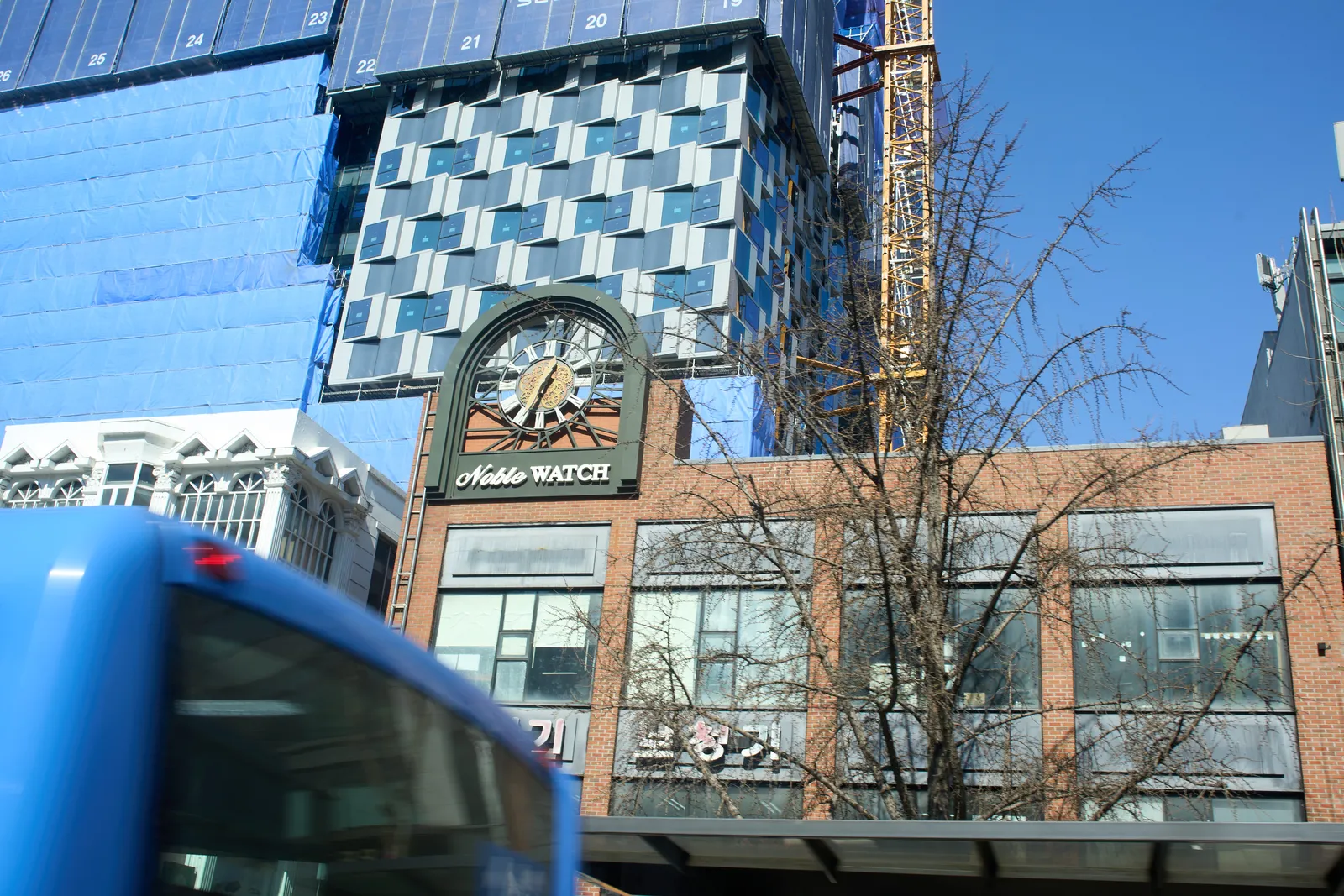 In the background there’s a large building. It is under construction. In front of this building there are two facades. A white, classical looking one, and one made from bricks. On top of this brick building there’s a rather large clock. It says Noble Watch beneath it. The clock points at 34 minutes past 12.