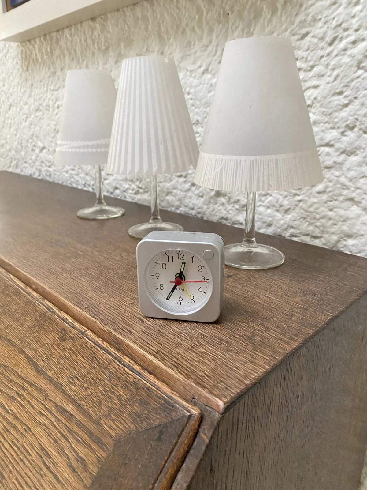 A small clock standing on a wooden cabinet. There are three white lamps behind it. The clock points at 26 minutes to one.