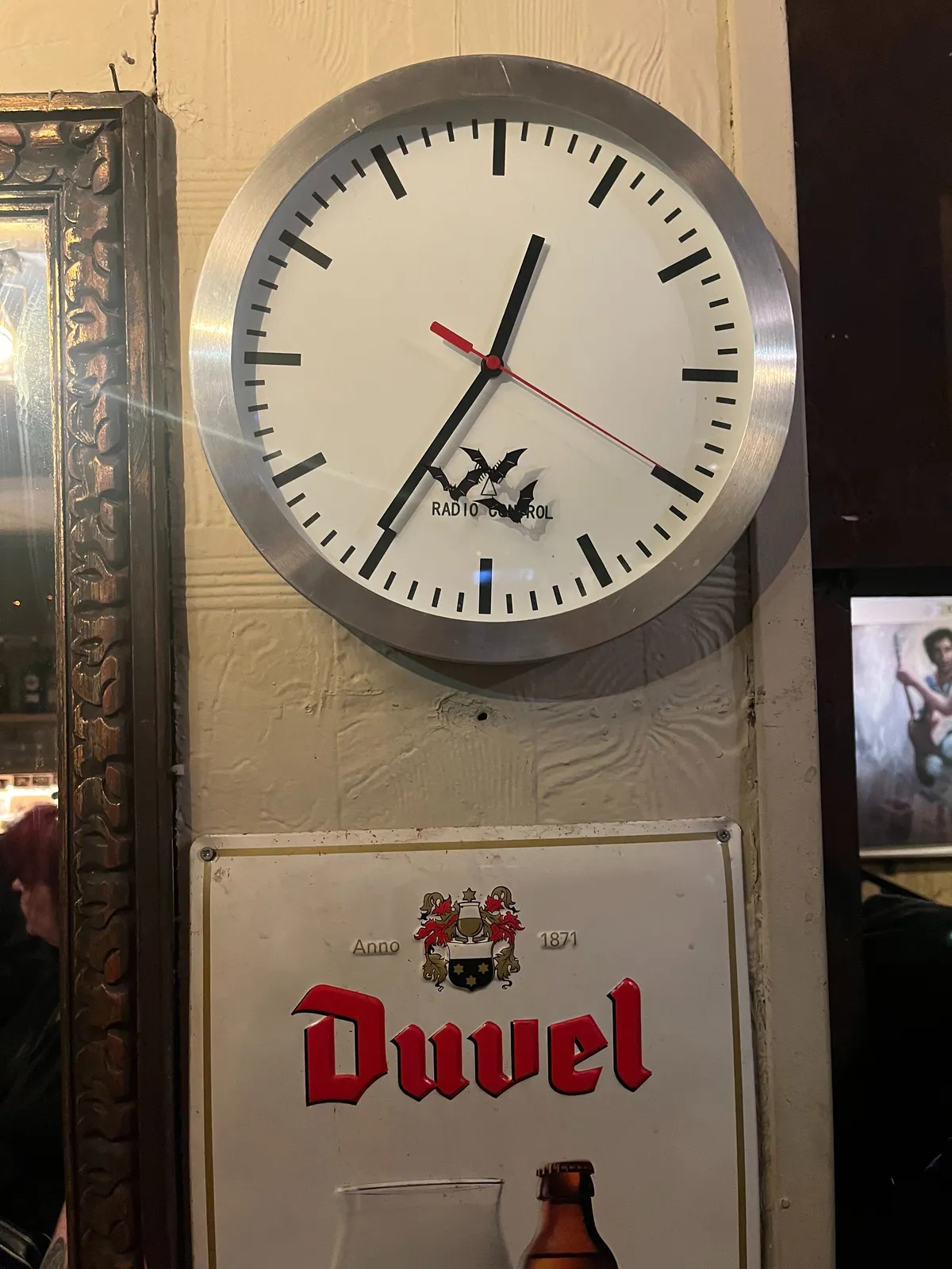A clock with three bats stickers on it. Below the clock there’s a sign that says Duvel. The clock points at 25 minutes to one.