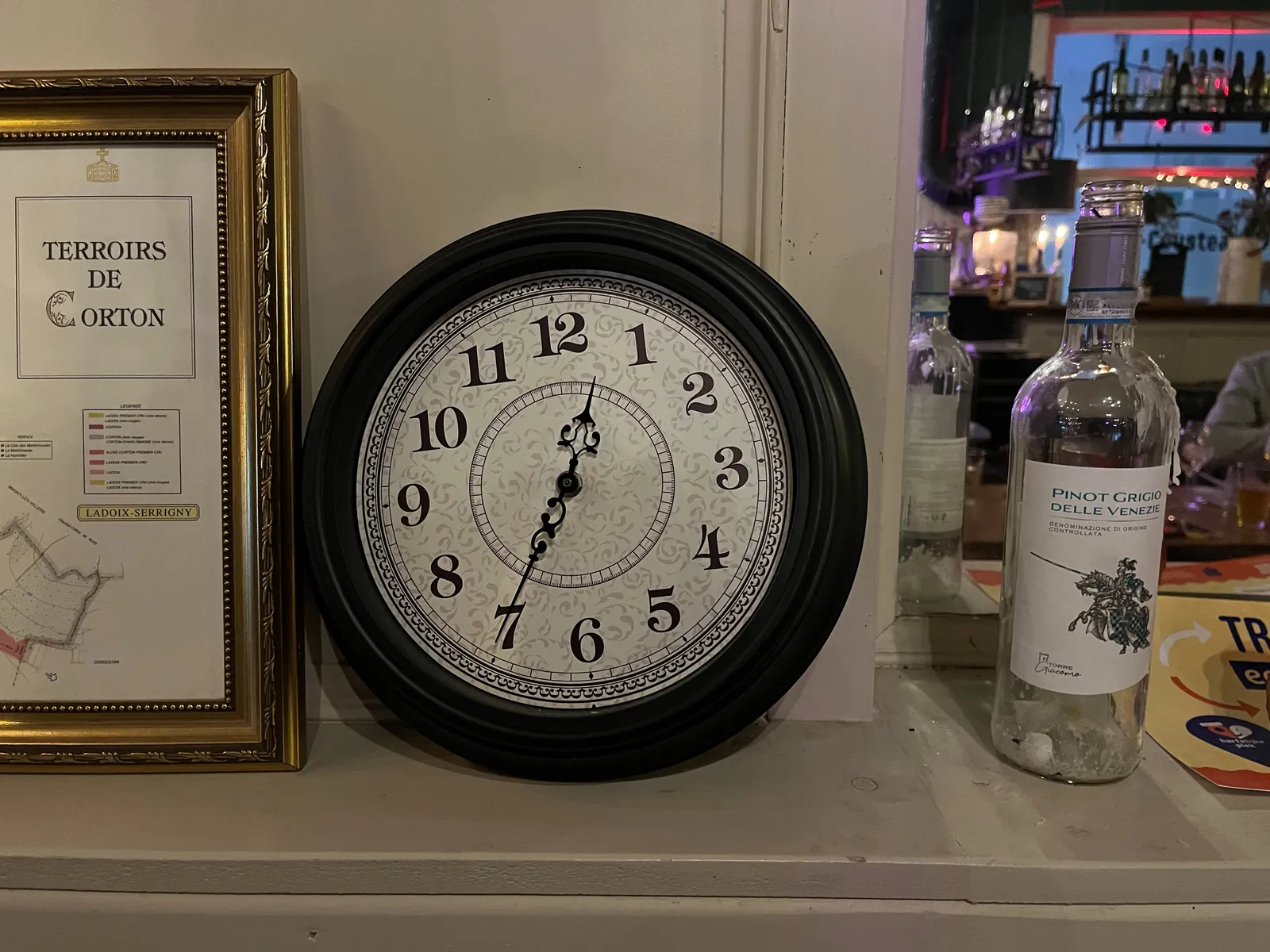 Next to an empty bottle of wine and a map of a vineyard in a golden frame, there’s a clock. It points at 25 minutes to one.