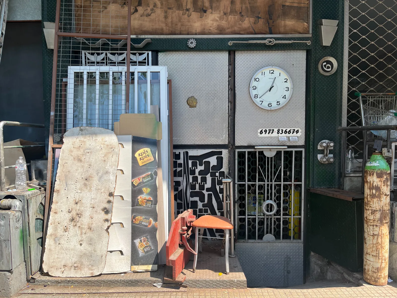Outside some sort of workshop, it’s a bit messy. There’s a clock attached to the door. On the clock it’s 22 minutes to one.